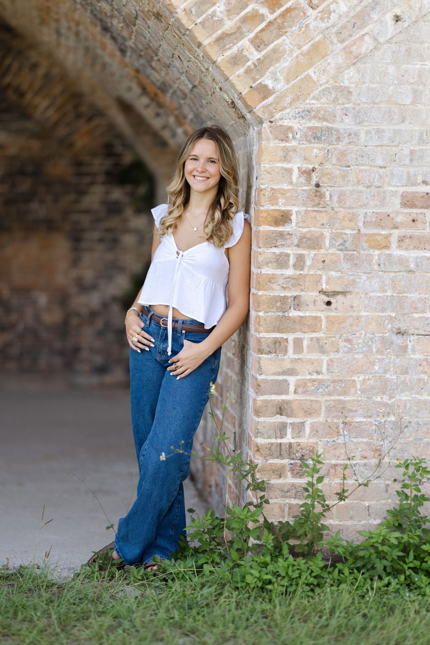 Casual lifestyle portrait against rustic brick archway with white top and blue jeans among green foliage.