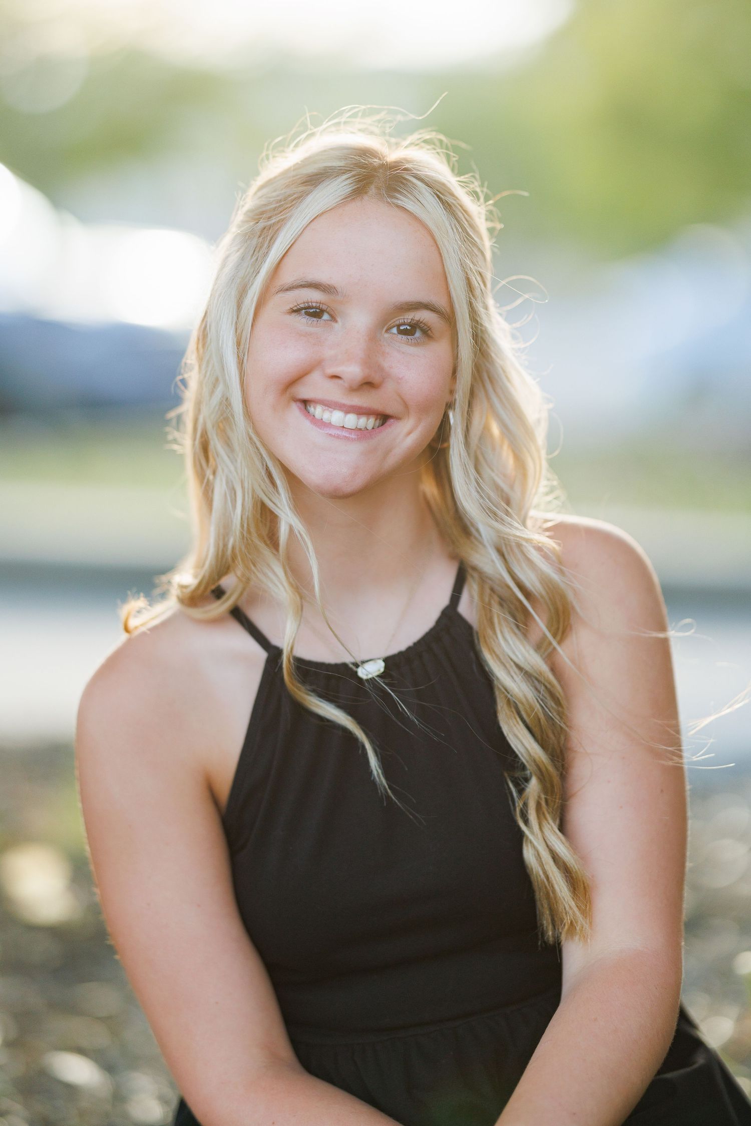 A young woman wearing a black halter dress poses outdoors with wavy blonde hair and a warm smile.