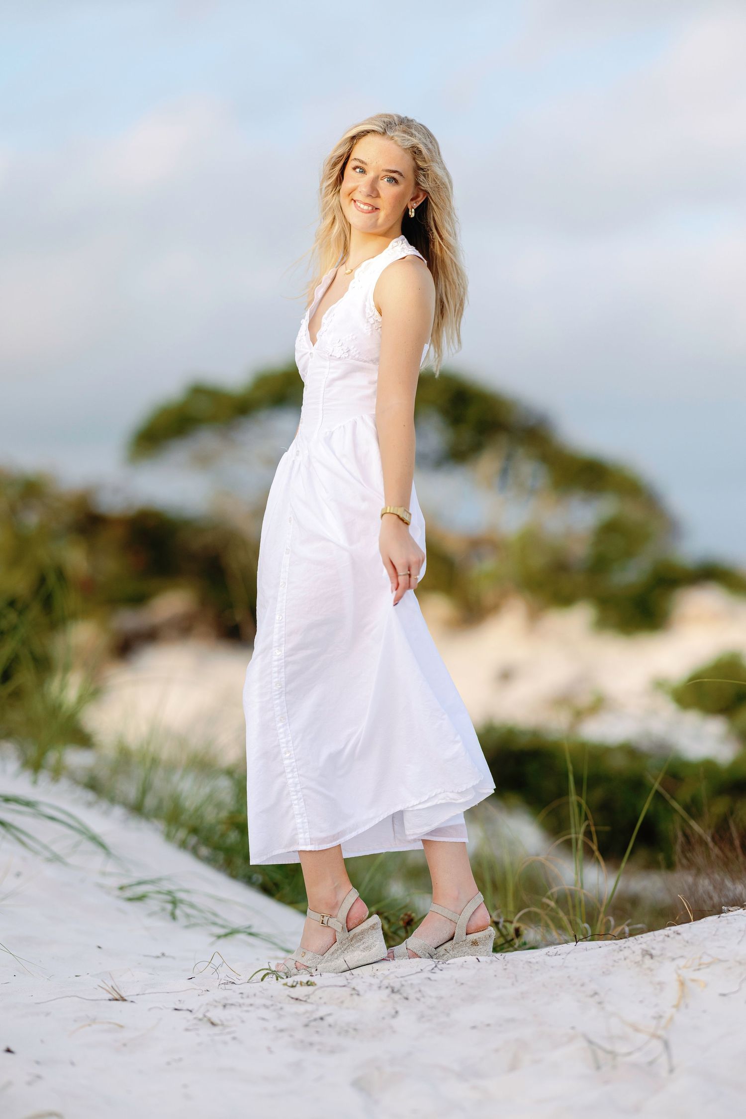 Person wearing flowing white dress stands on sandy dunes against coastal backdrop.