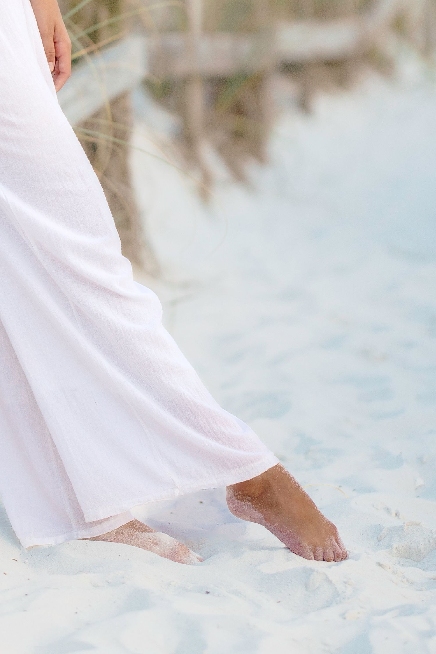 Bare feet walking on white sandy beach with flowing white dress.