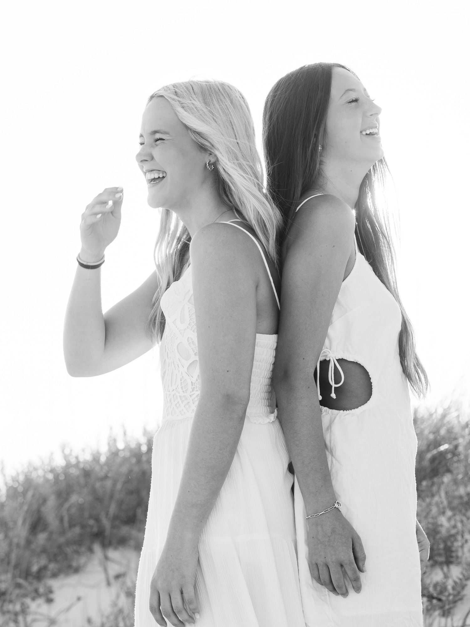Black and white photo of two friends laughing back to back on beach.