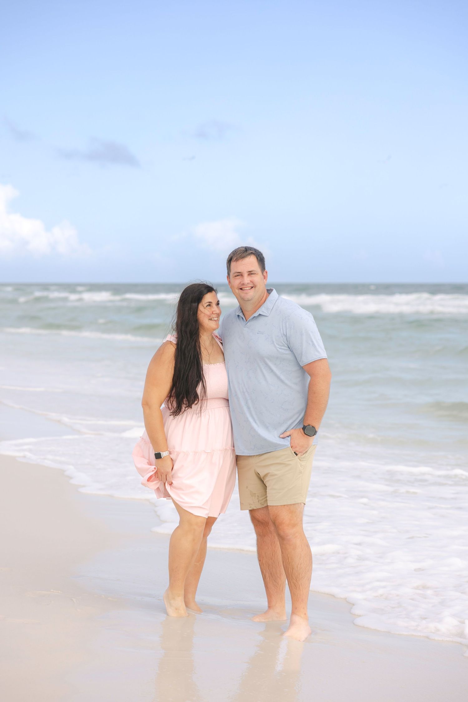Couple in casual beachwear shares romantic moment along shoreline with waves crashing behind them.