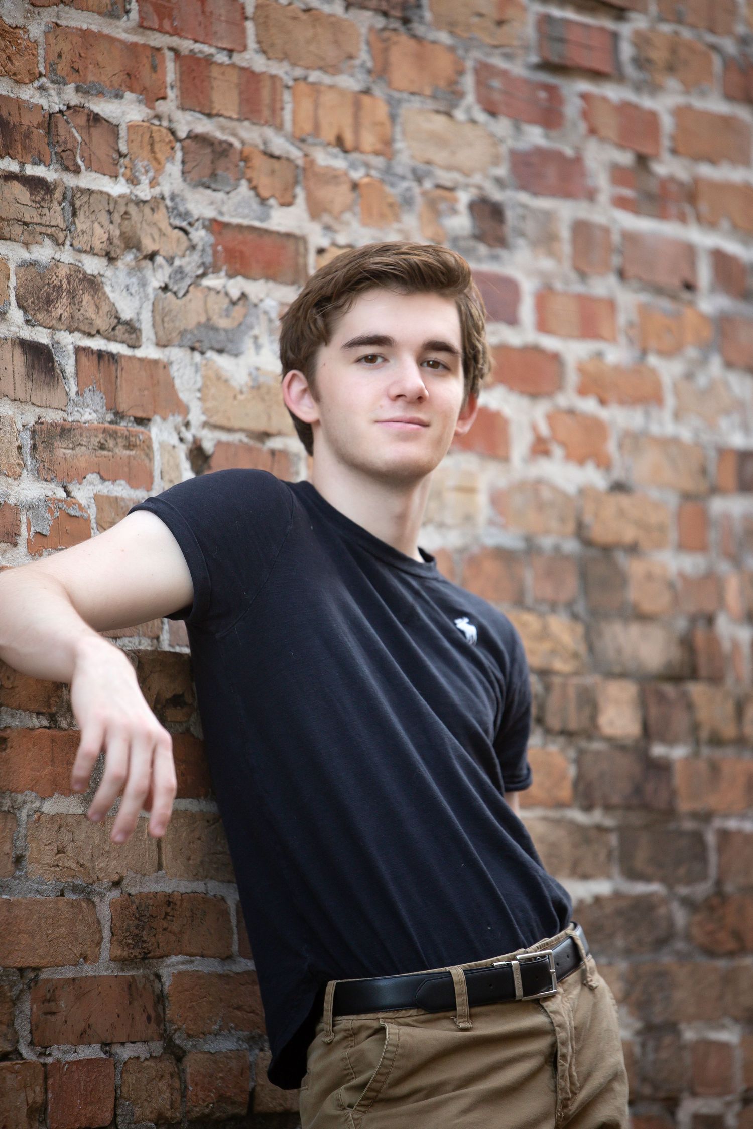 Young man in black t-shirt leaning against rustic brick wall, smiling confidently in casual outdoor portrait.