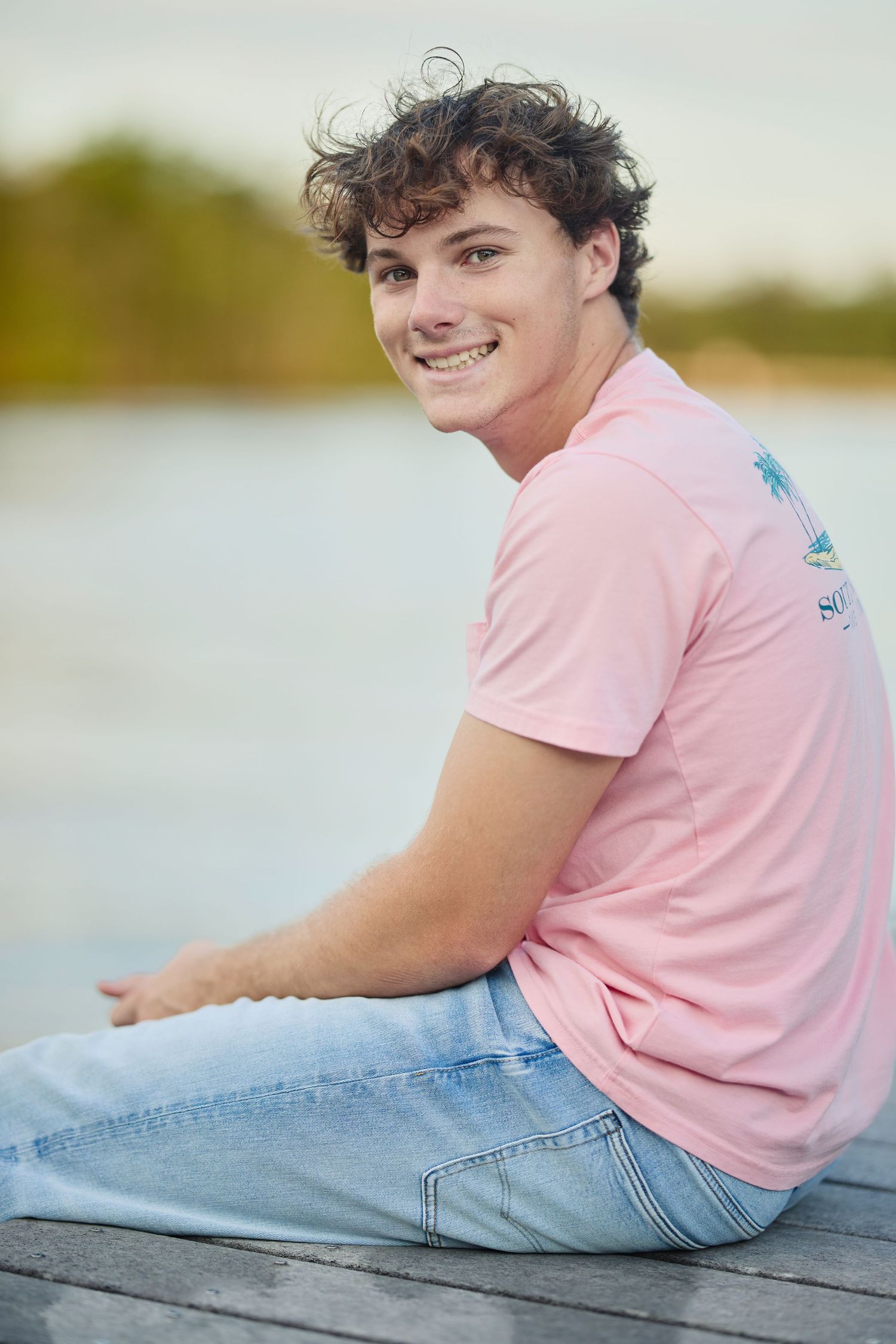Smiling young man with curly hair sitting on a dock in a pink t-shirt and light jeans.