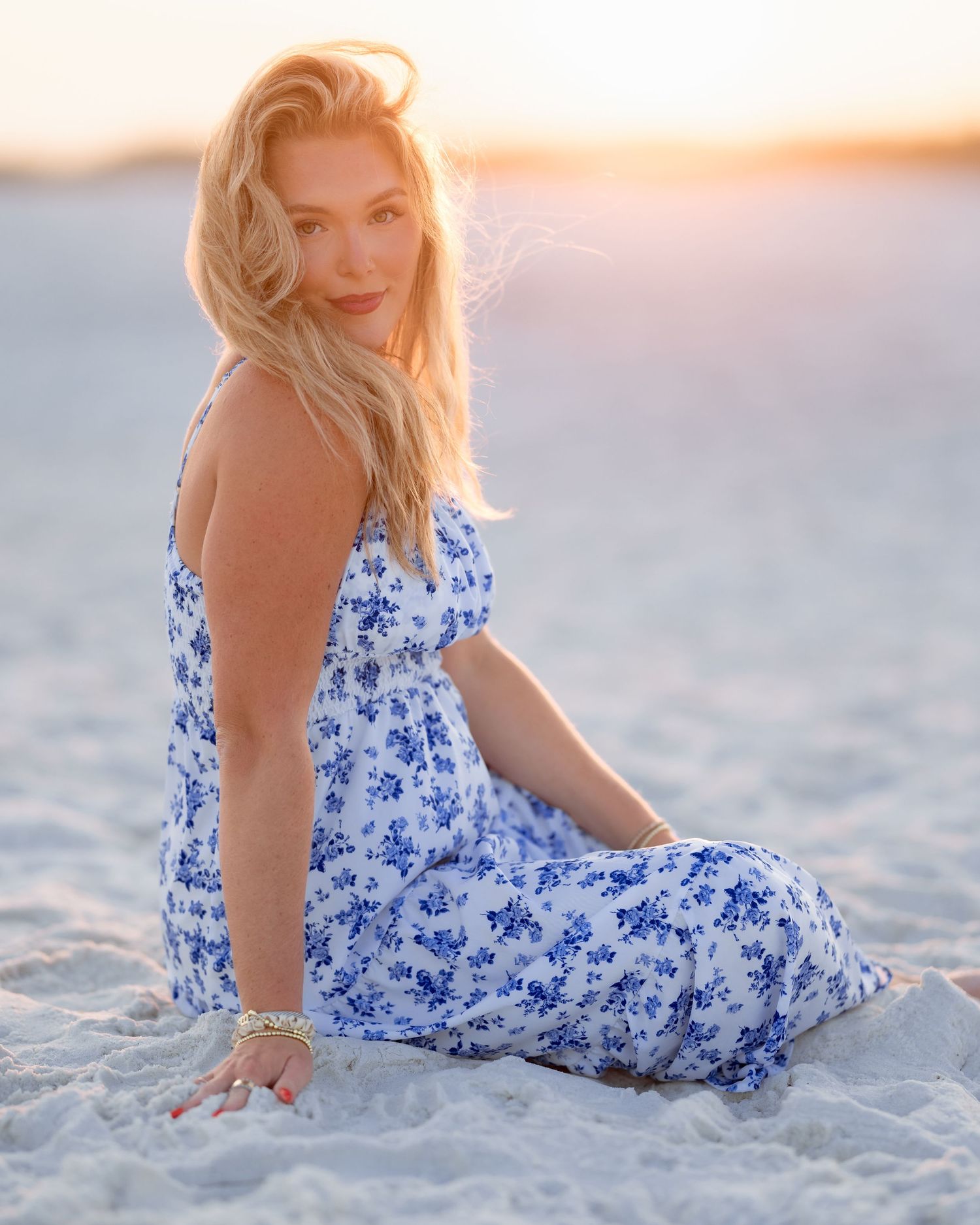 Woman in blue floral dress sitting on sandy beach at golden hour sunset.