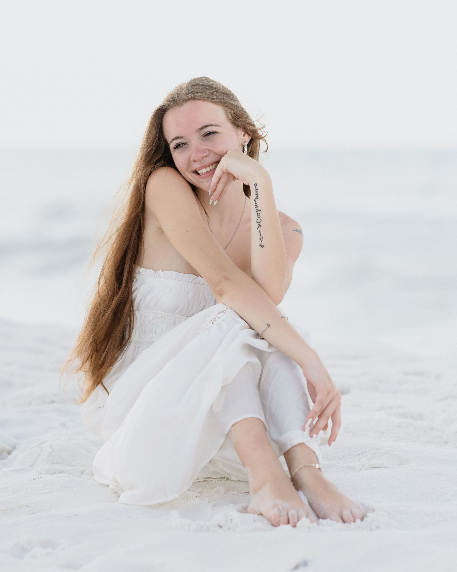 Young woman with long hair sits smiling on white sand beach, wearing white dress and pants, barefoot.