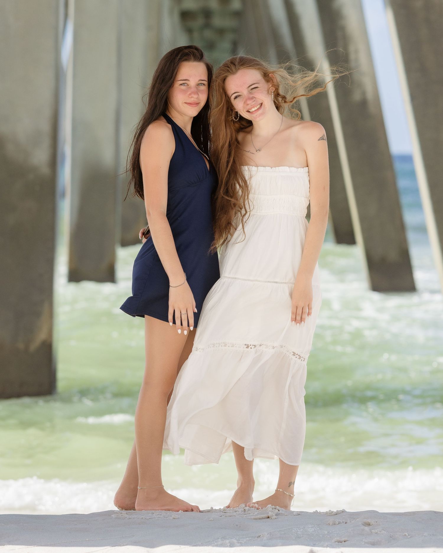 Two young women posing together on a beach, one in navy dress and one in white maxi dress, with pier in background.