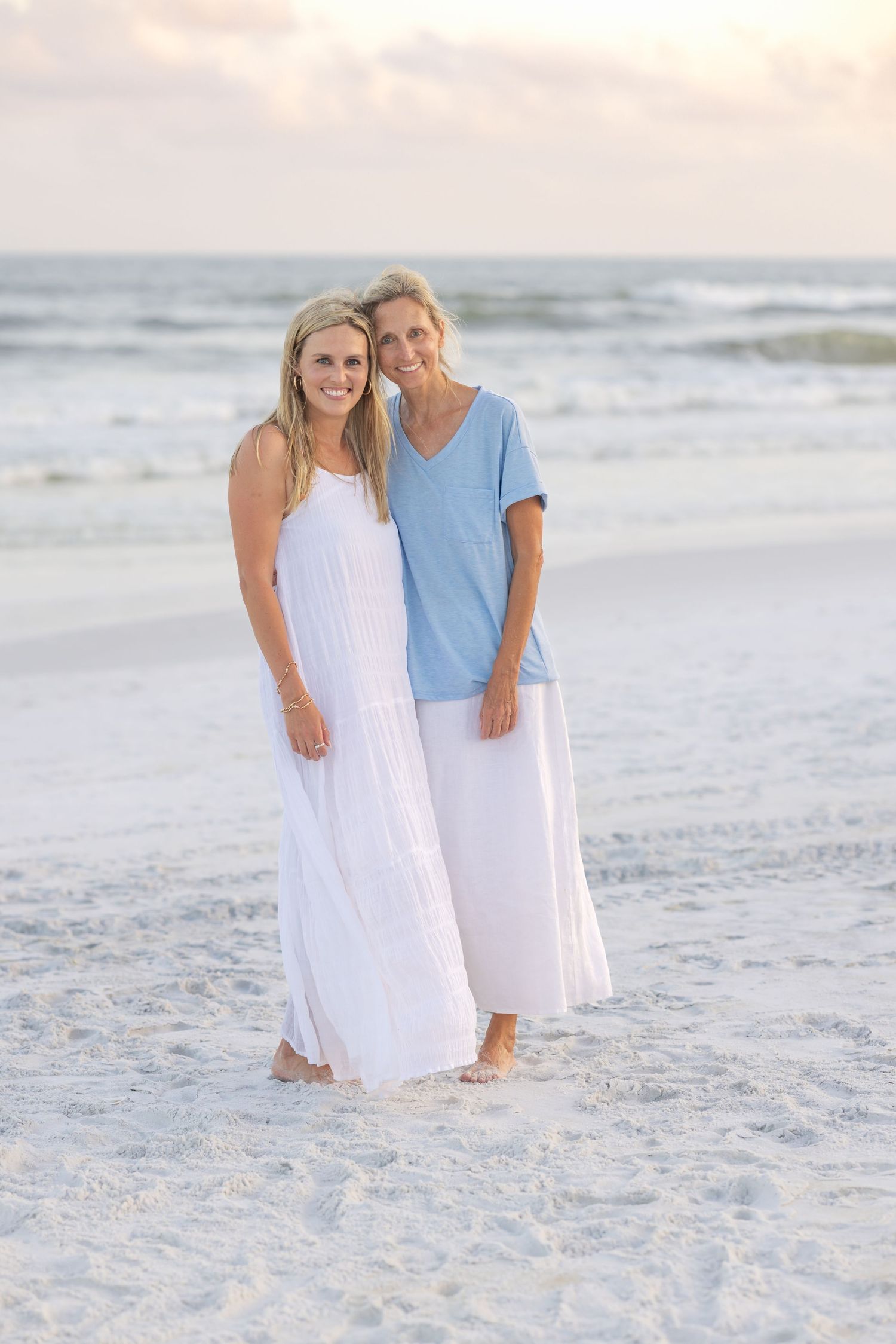 Two blonde women in white outfits smiling together on a sandy beach at sunset.