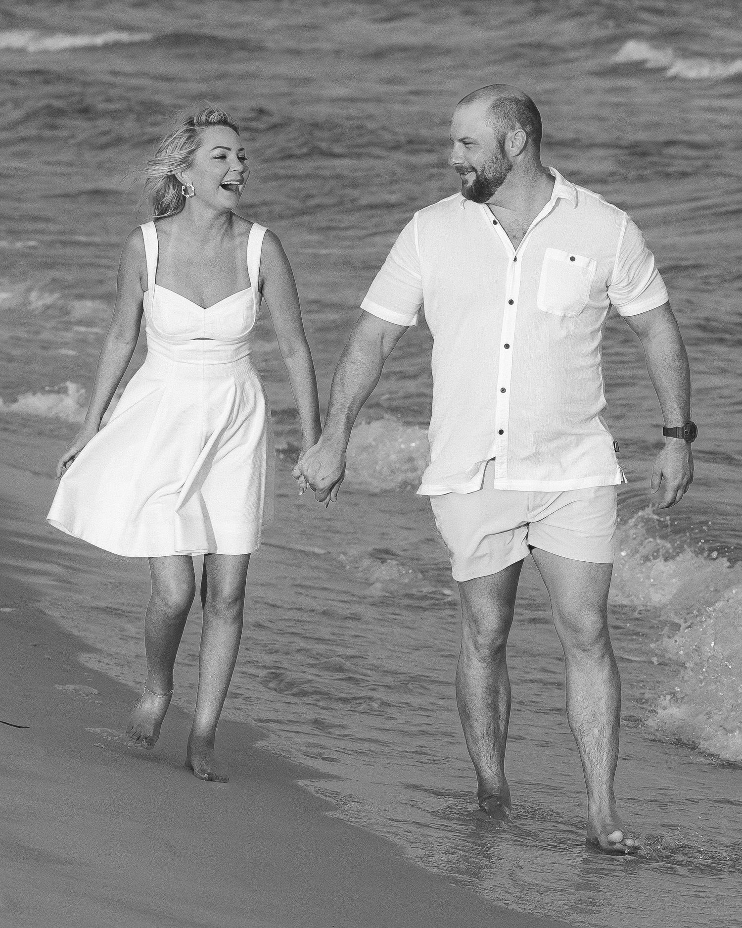 Couple in white outfits walk hand in hand laughing along ocean shoreline in black and white photo.
