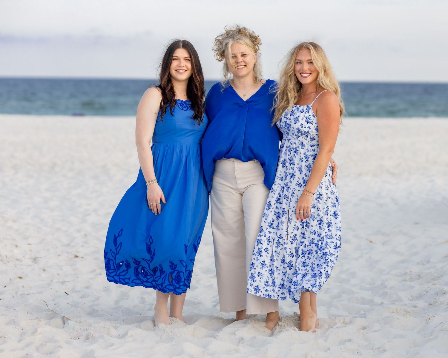 Three women in blue outfits smiling together on a white sand beach on a sunny day.
