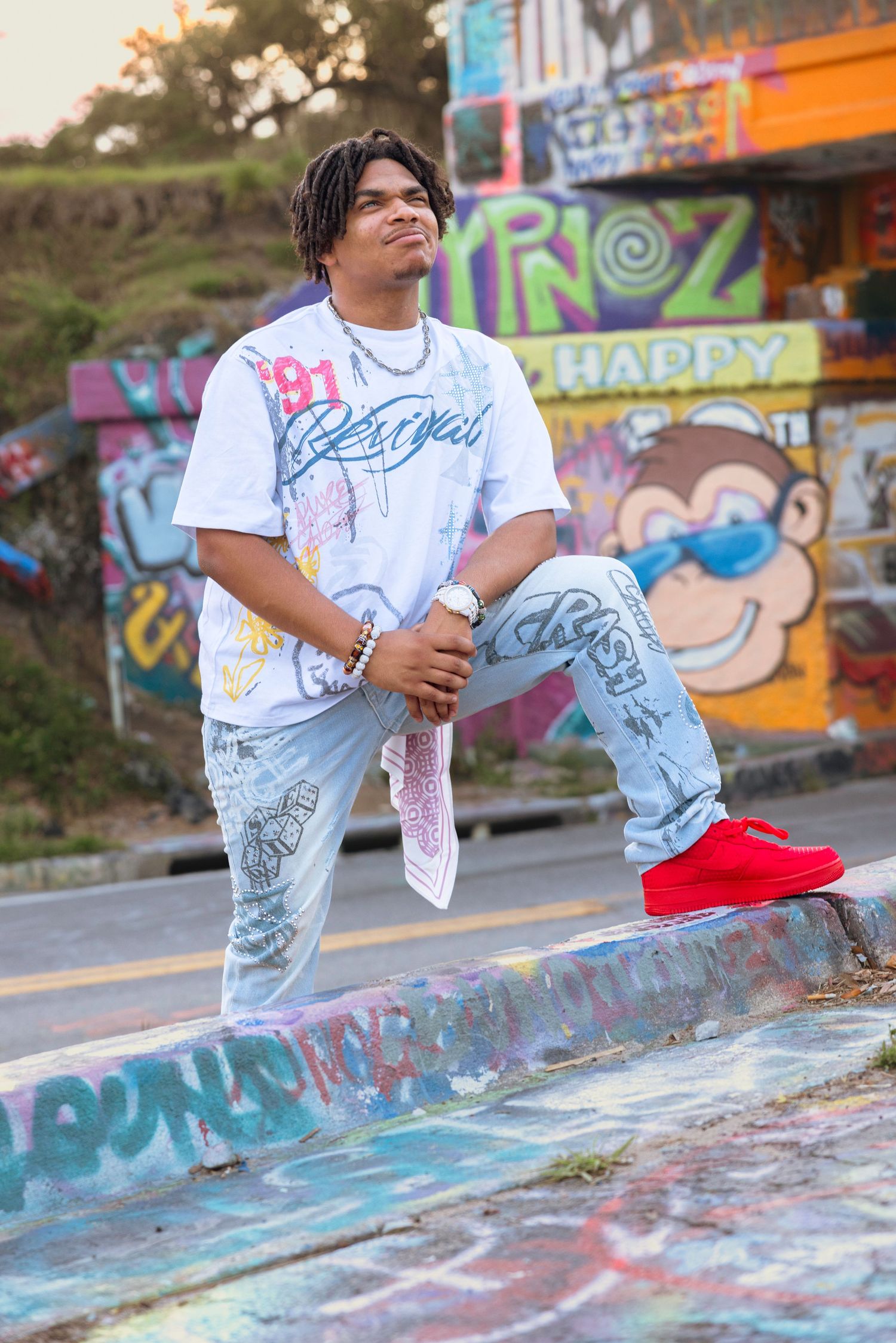 Young man in white graphic tee, light jeans, and red sneakers poses at colorful graffiti skatepark.