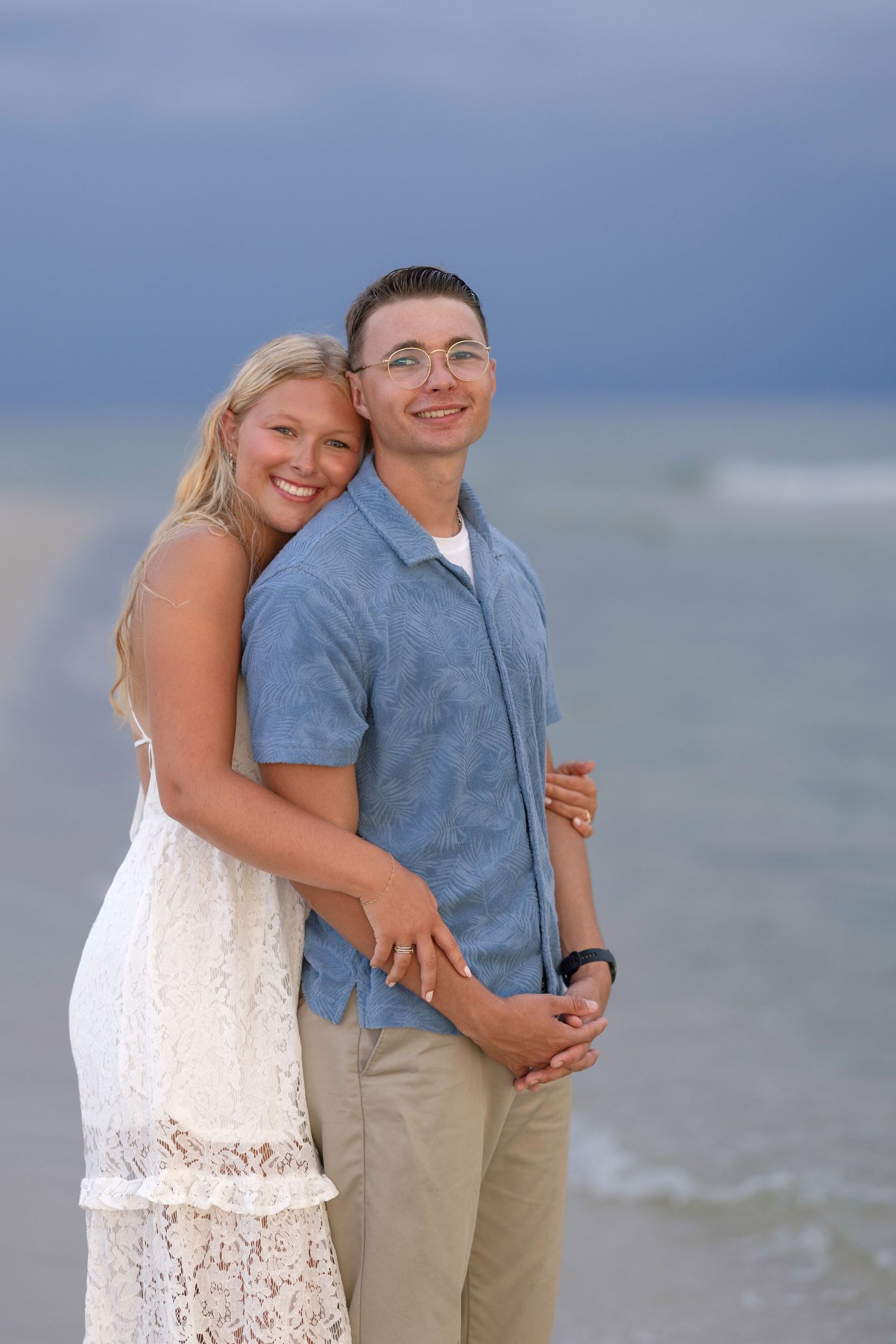 Happy couple embracing on a beach, woman in white lace dress and man in blue shirt, with ocean waves behind them.