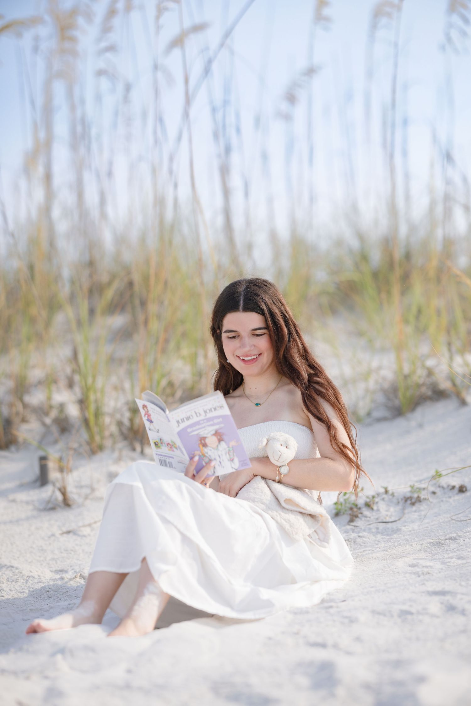 Young woman in white dress reading book while sitting on sandy beach dunes with sea oats.