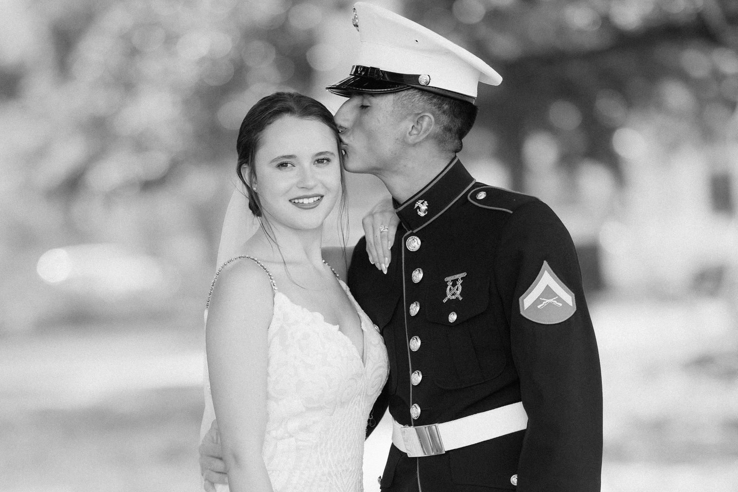 Black and white wedding photo of a Marine in dress uniform kissing bride on cheek outdoors.