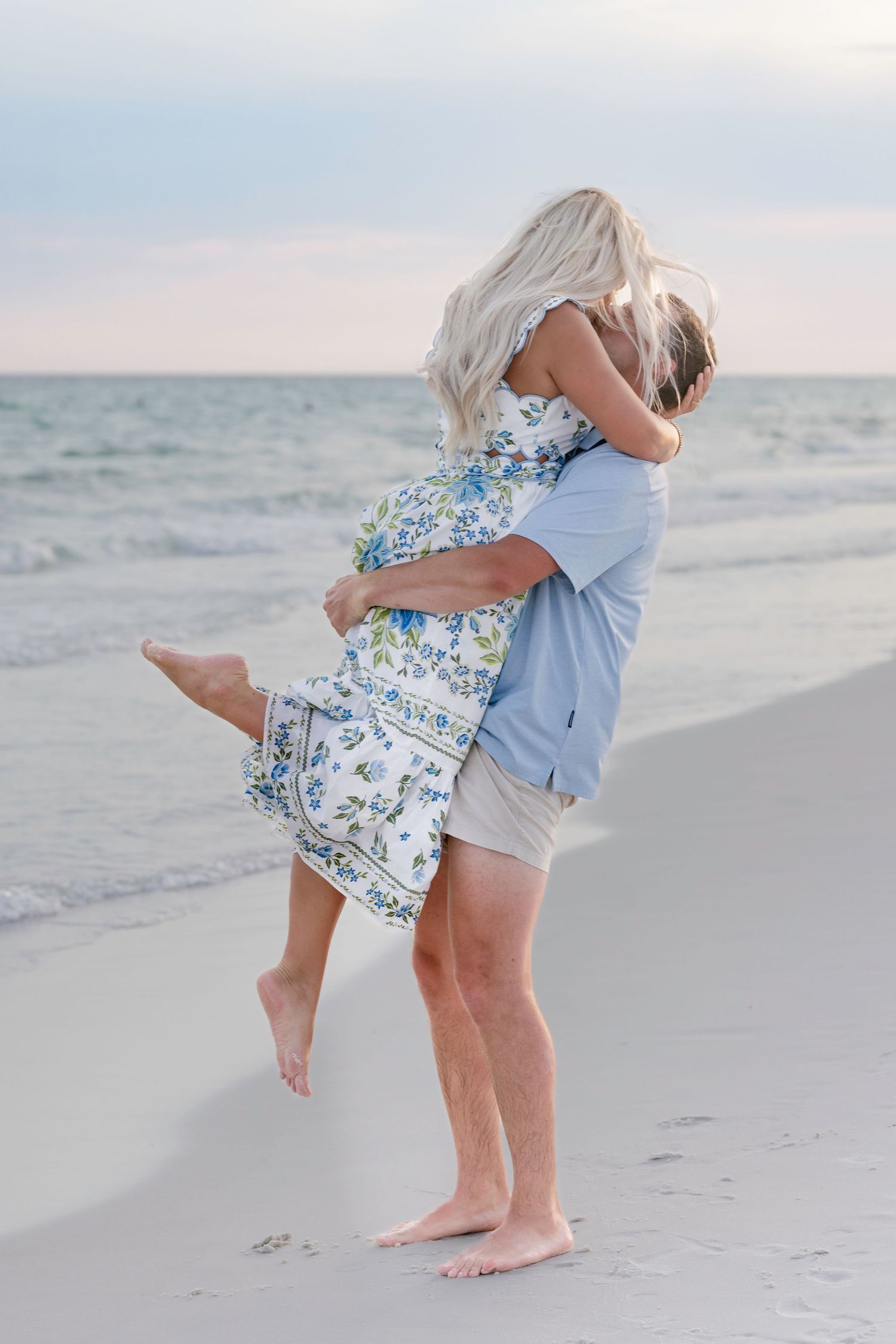 Couple sharing a romantic embrace on a sandy beach at sunset, woman in floral dress lifted by man.