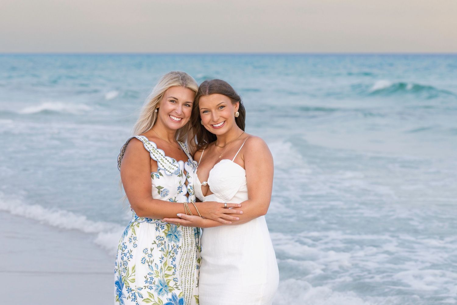 Two women smiling and embracing on a sandy beach with ocean waves, wearing white and floral dresses.