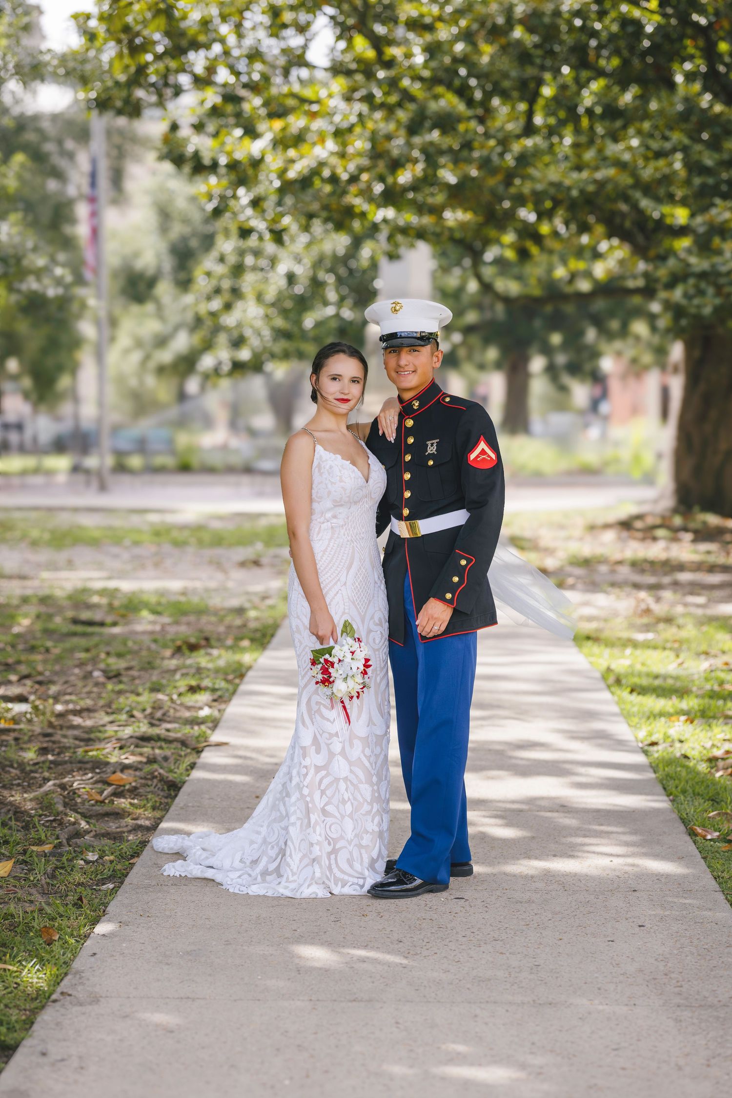 Marine in dress blues and bride in white lace gown pose together on a sunny sidewalk holding a bouquet.