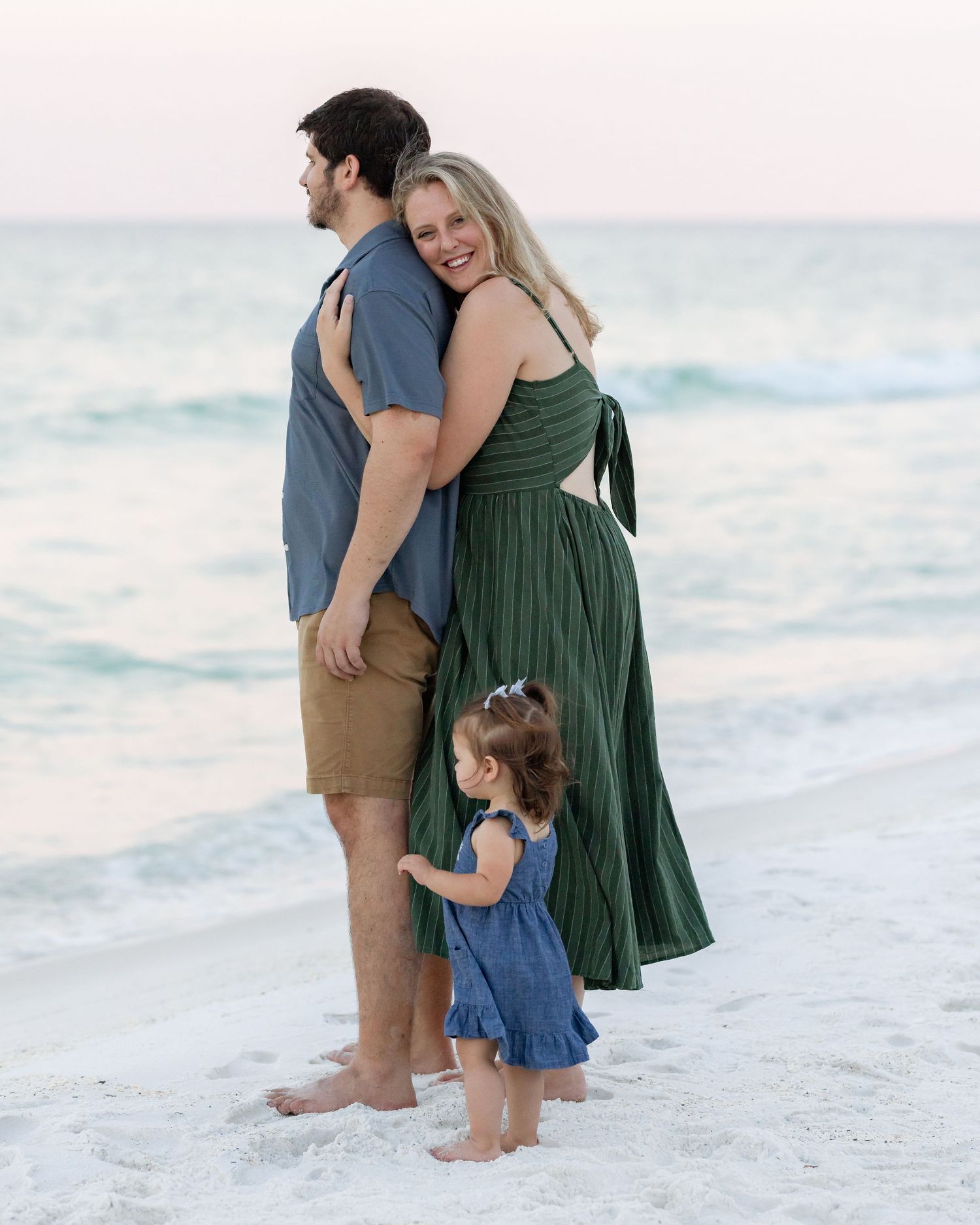 Family of three poses on a sandy beach at sunset, parents embracing while toddler girl stands between them.