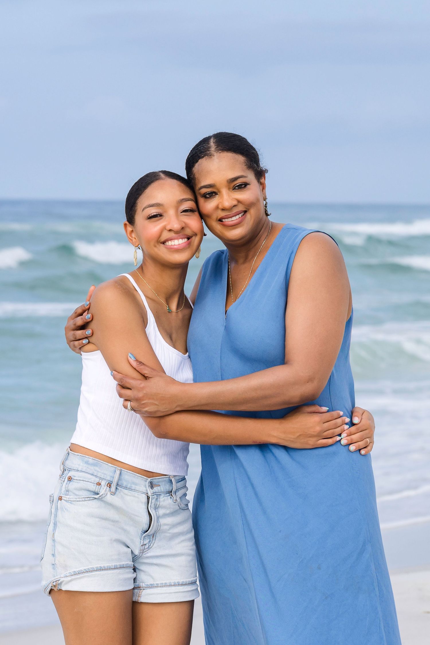 Two women embracing and smiling together on a beach with ocean waves in the background.