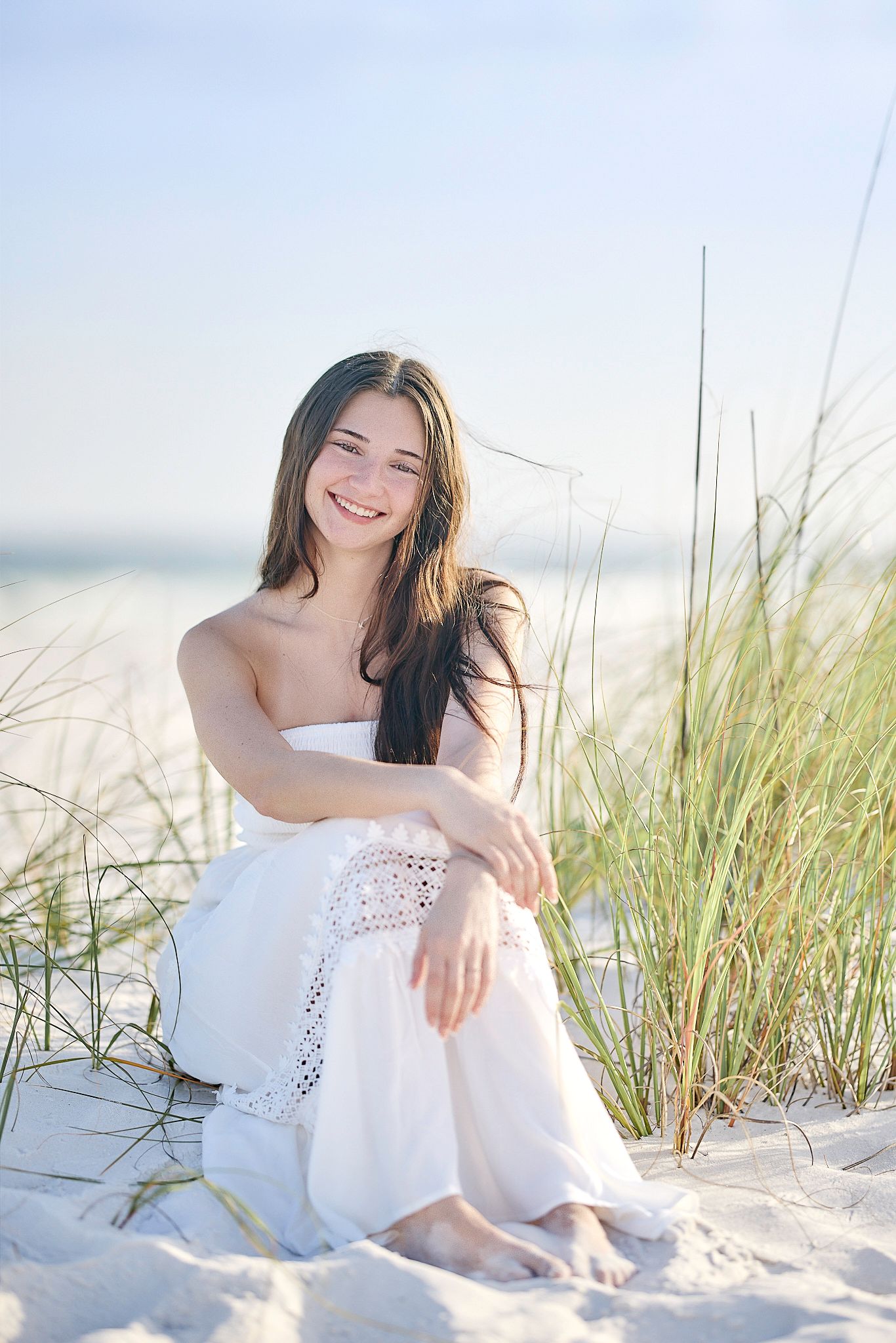Young woman in white dress sitting on sandy beach dunes, smiling with long dark hair, beach grass behind her.