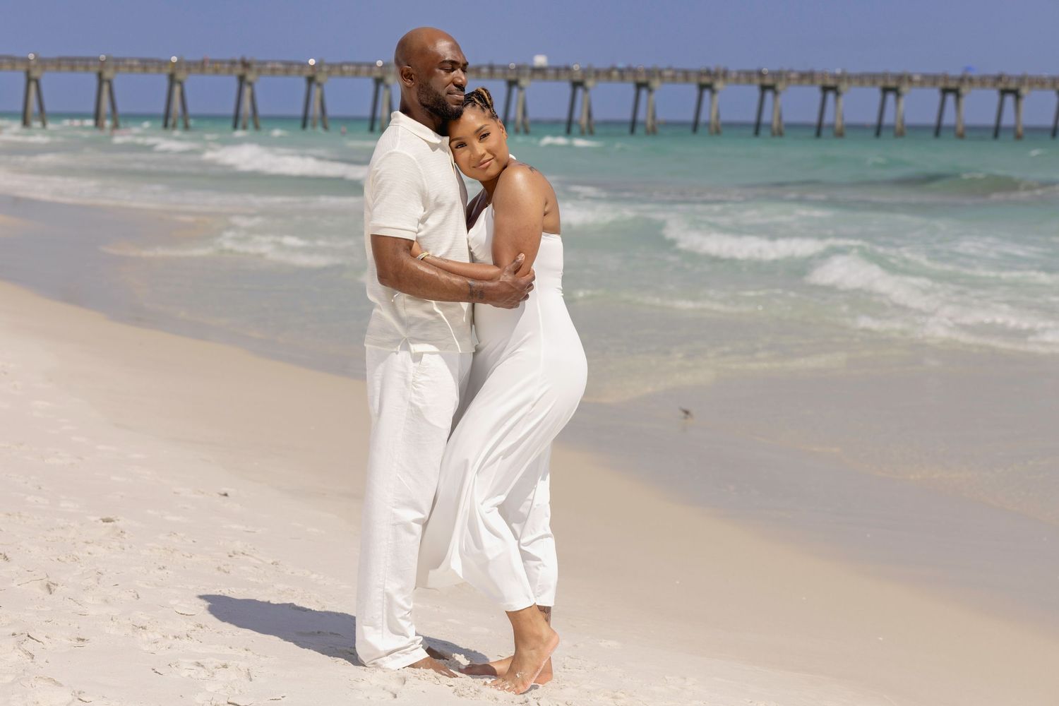Couple in white outfits embracing on a sandy beach with turquoise ocean waves and a pier in the background.