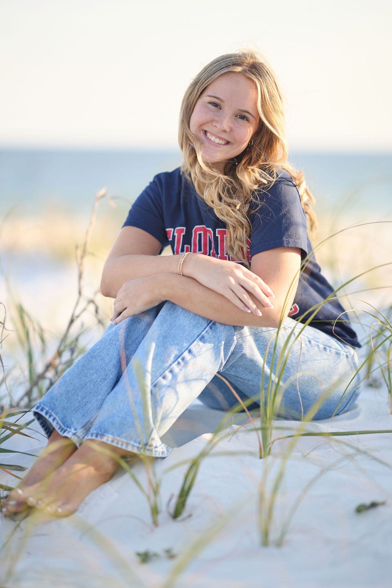 Smiling blonde teen girl sitting on white sand beach dunes wearing jeans and a navy Florida shirt.