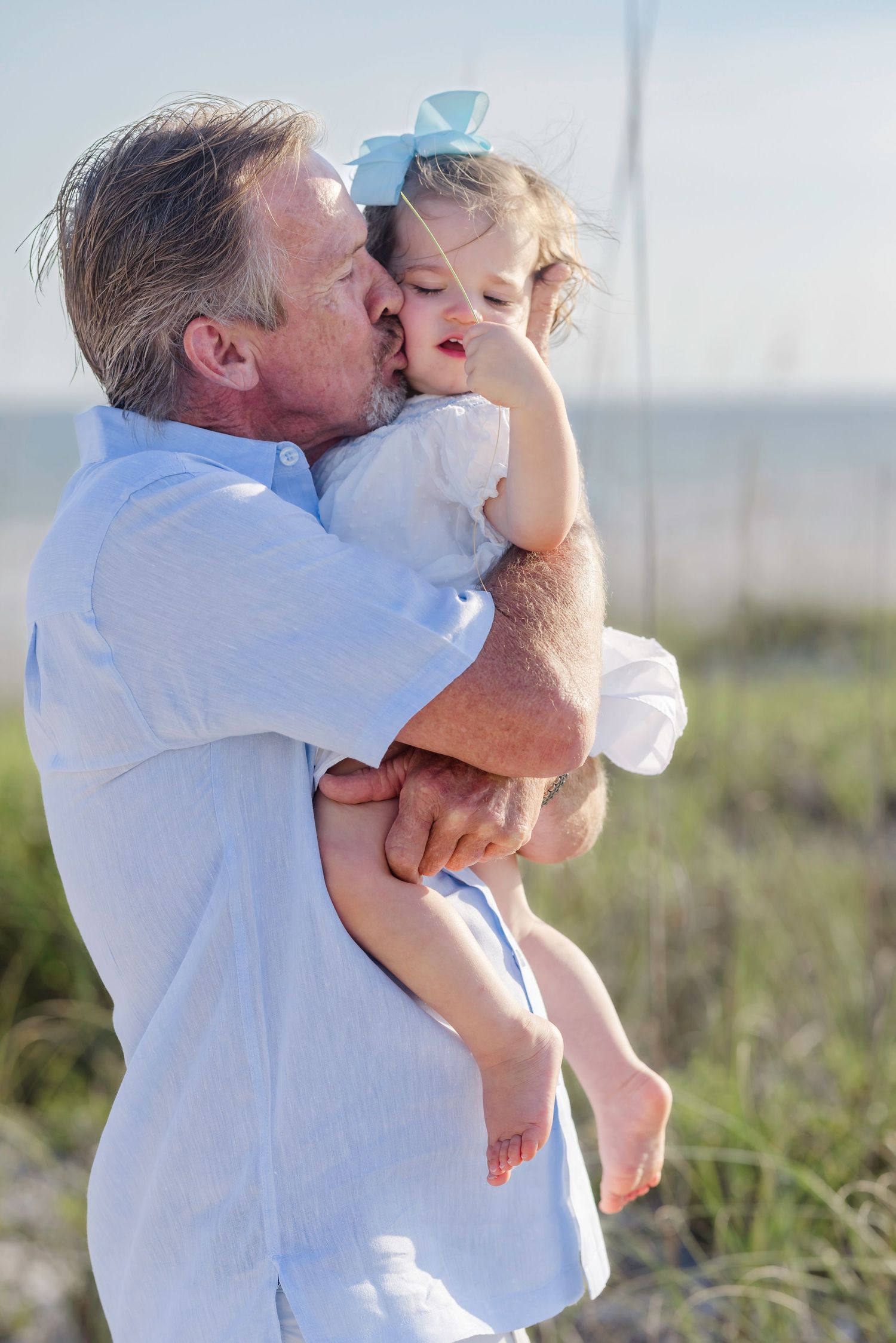 Grandfather kissing toddler granddaughter with blue bow at sunny beach.