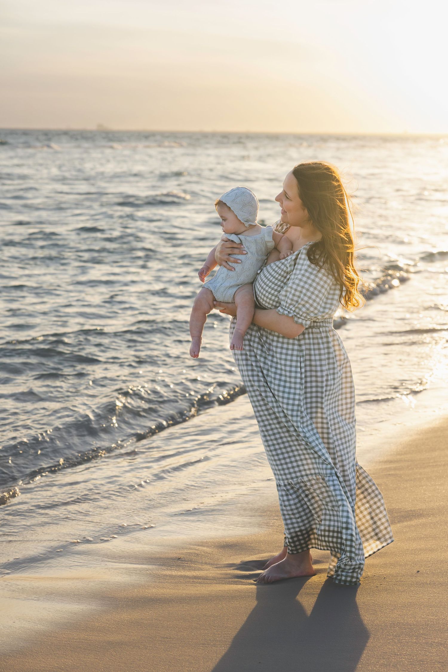 A mother in a plaid dress holds her baby on a sandy beach at golden hour sunset.