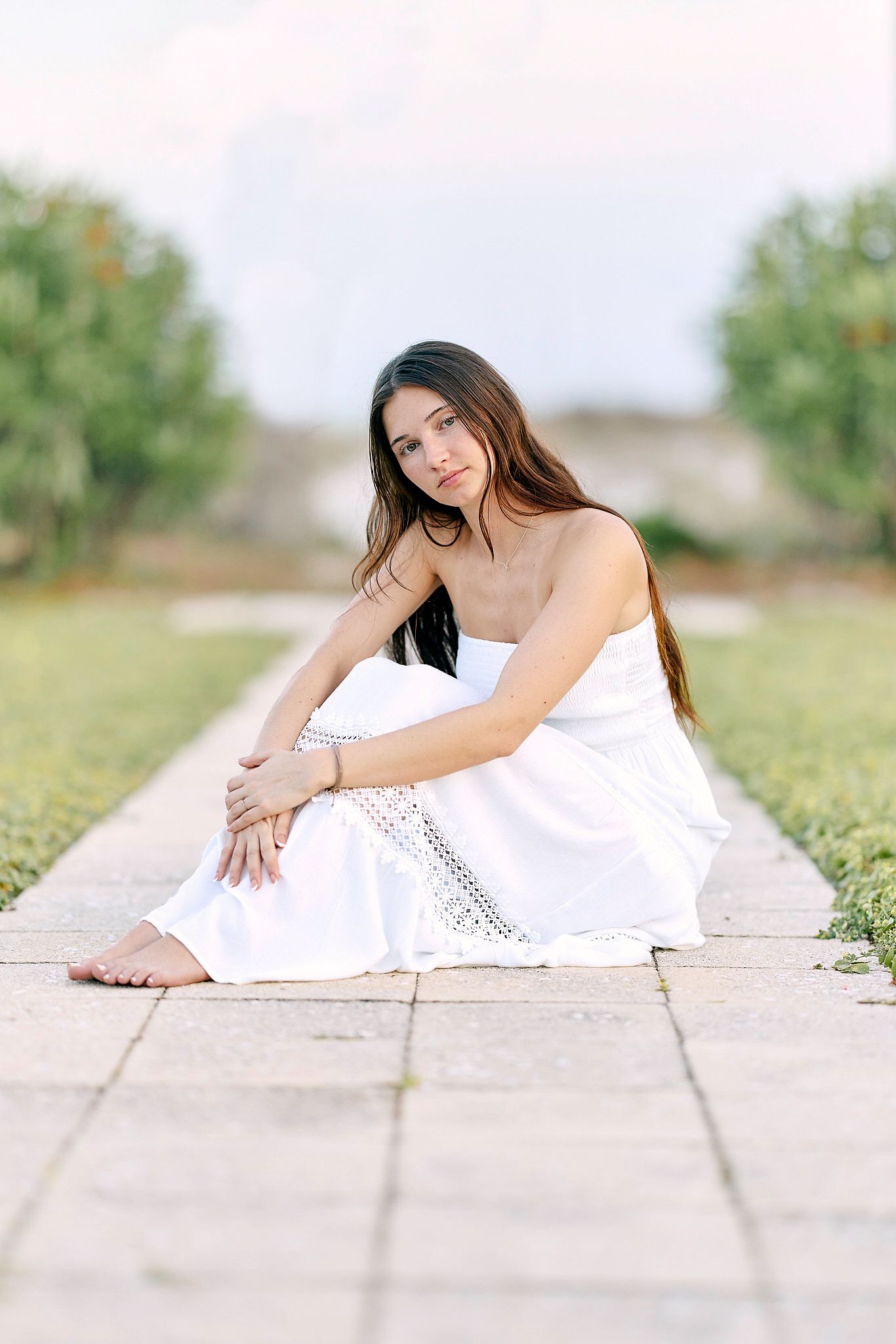 Young woman in white strapless dress sitting barefoot on stone path in lush green garden, gazing at camera.