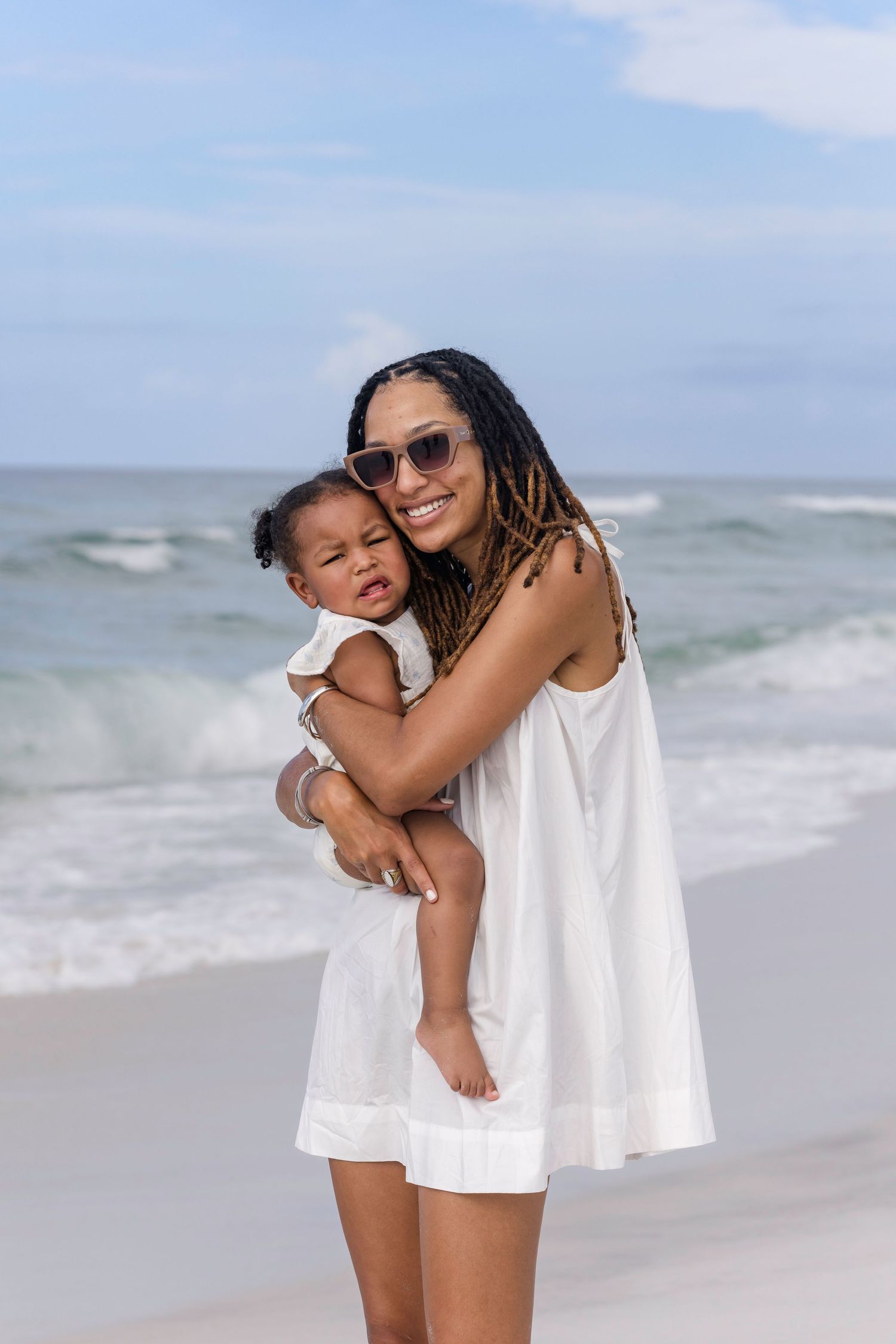 Smiling Black mother in white dress holds her baby girl while standing on a sunny beach with ocean waves behind them.