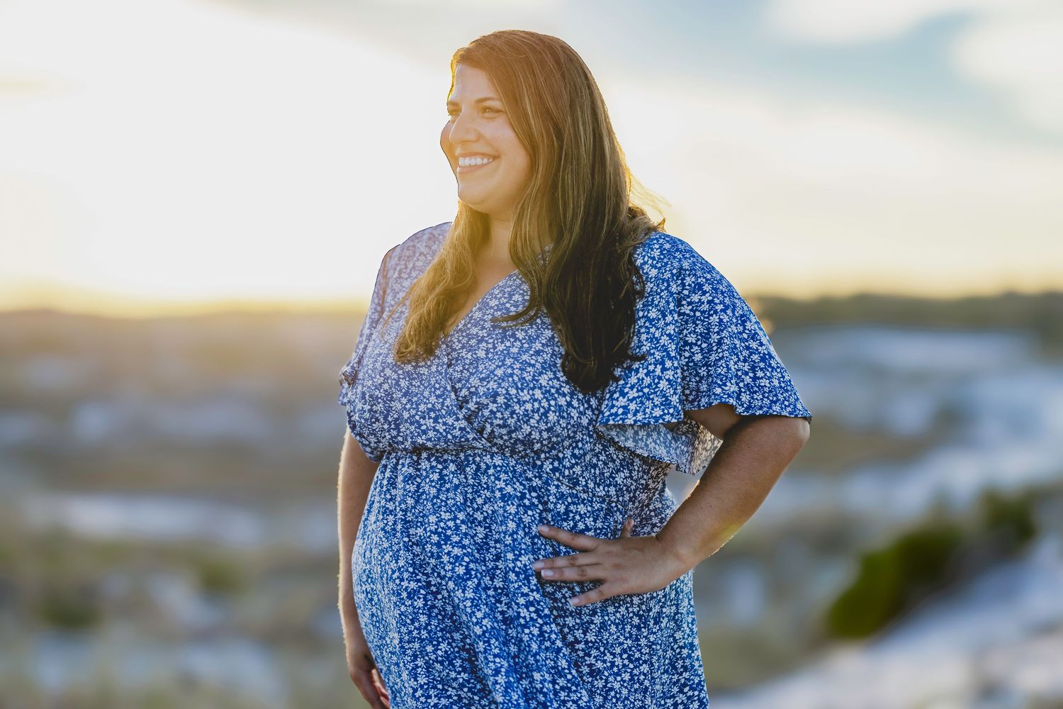Smiling pregnant woman in blue floral dress holding her belly at sunset outdoors.