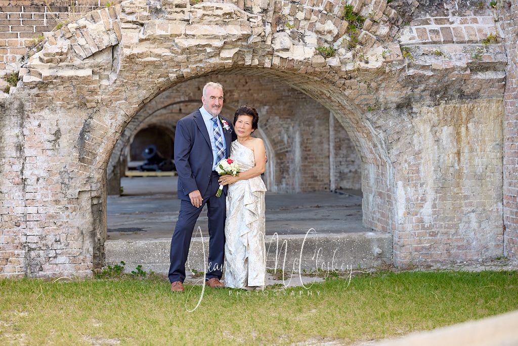 A couple poses under a stone archway at Fort Pickens, with weathered walls and green grass in the foreground.