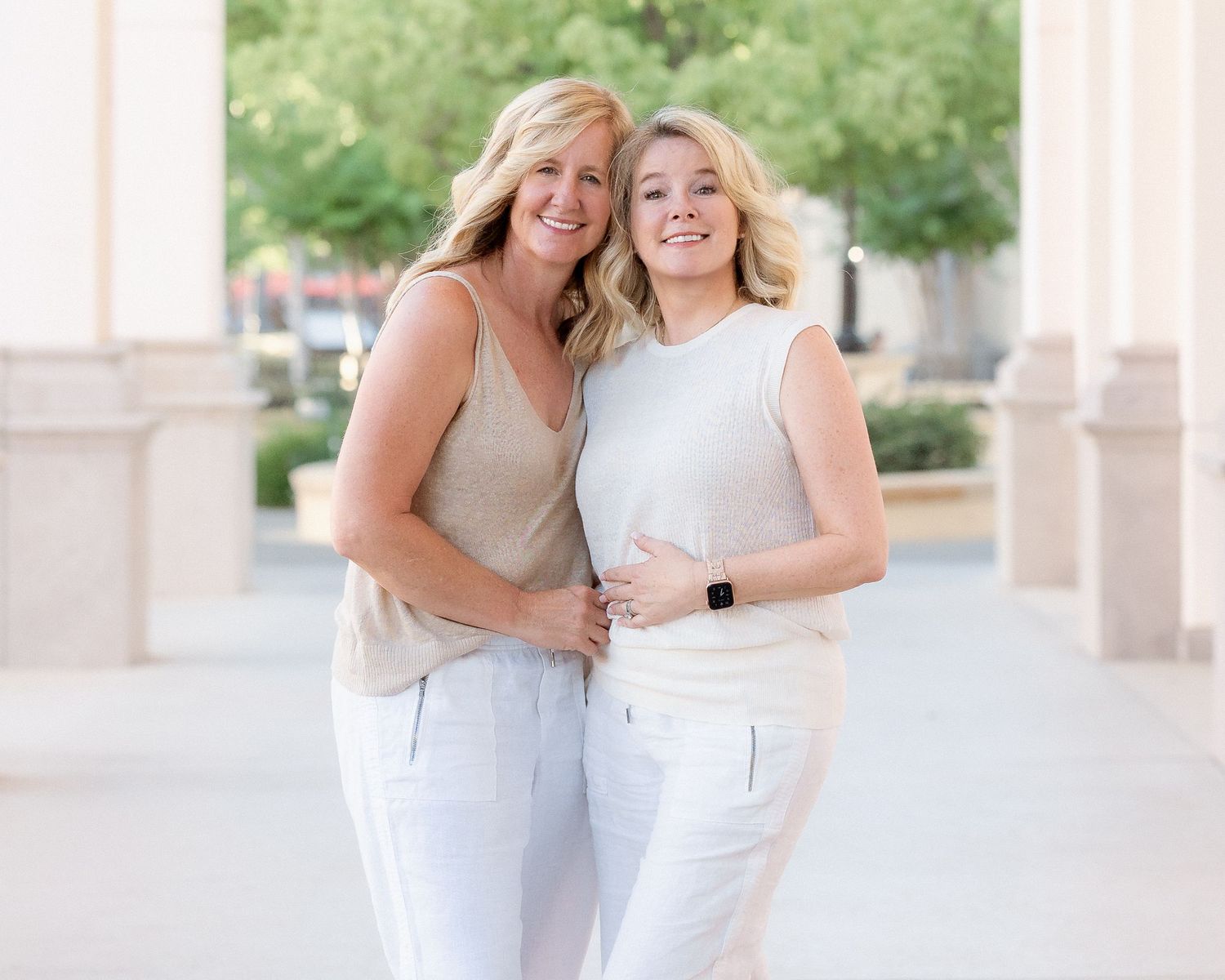 Two smiling blonde women in neutral outfits pose together outdoors near columns on a sunny day.