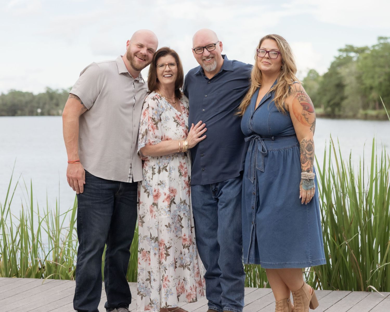 Four adults smiling together on a wooden dock by a lake, posing for a family portrait outdoors.