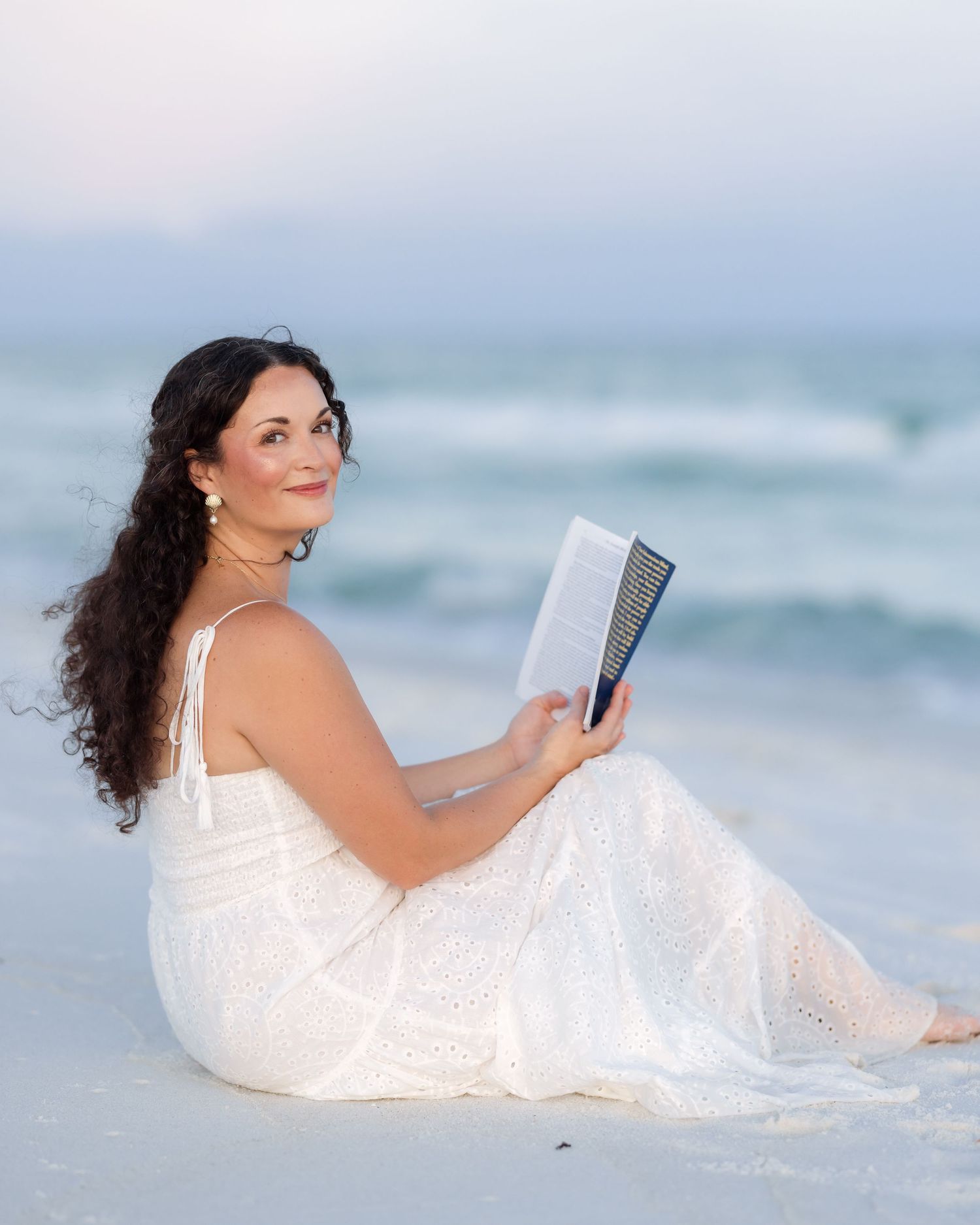 Woman in white dress sitting on beach sand reading a book.