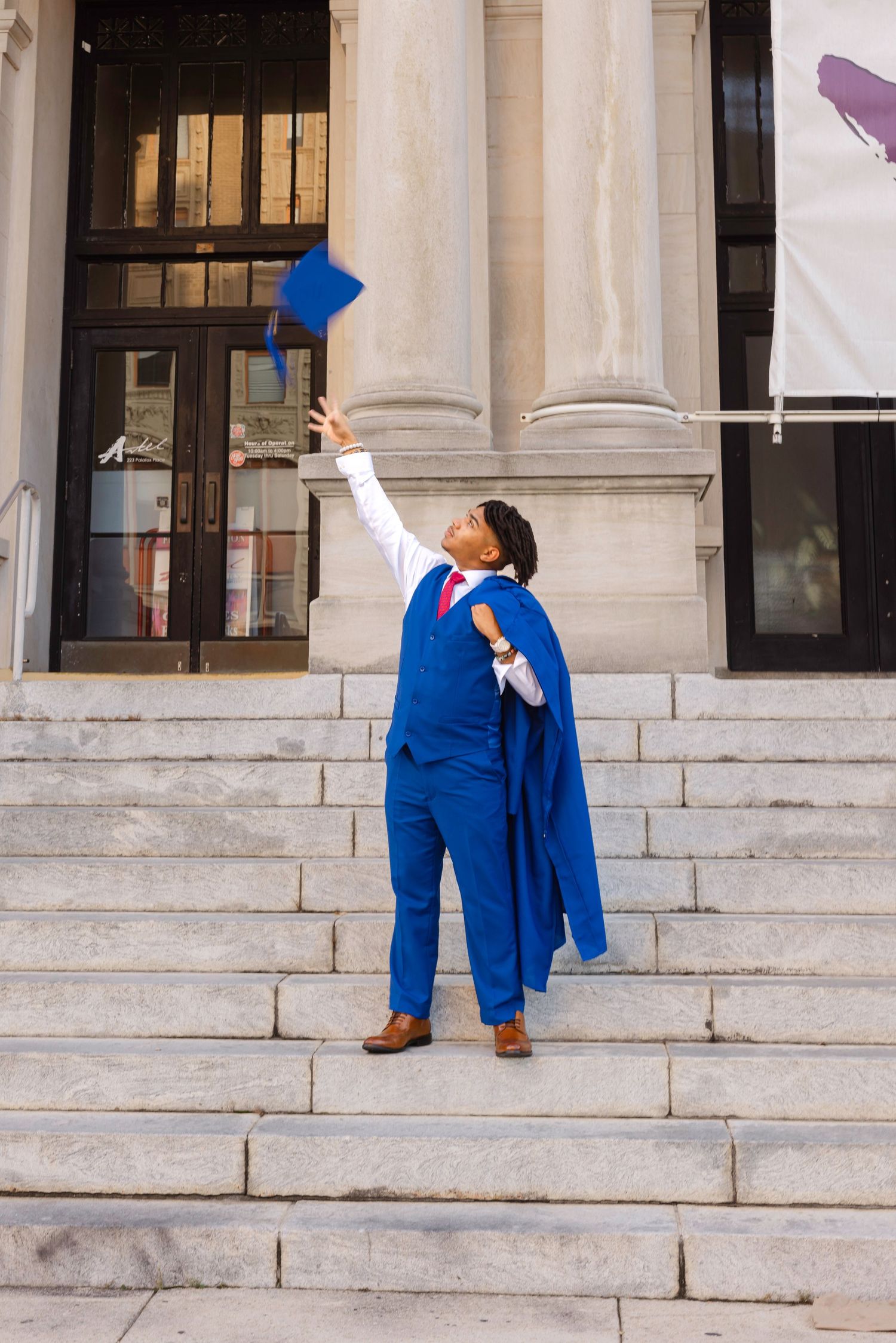 Graduate in blue gown and suit tosses cap on university steps, celebrating academic achievement outdoors.