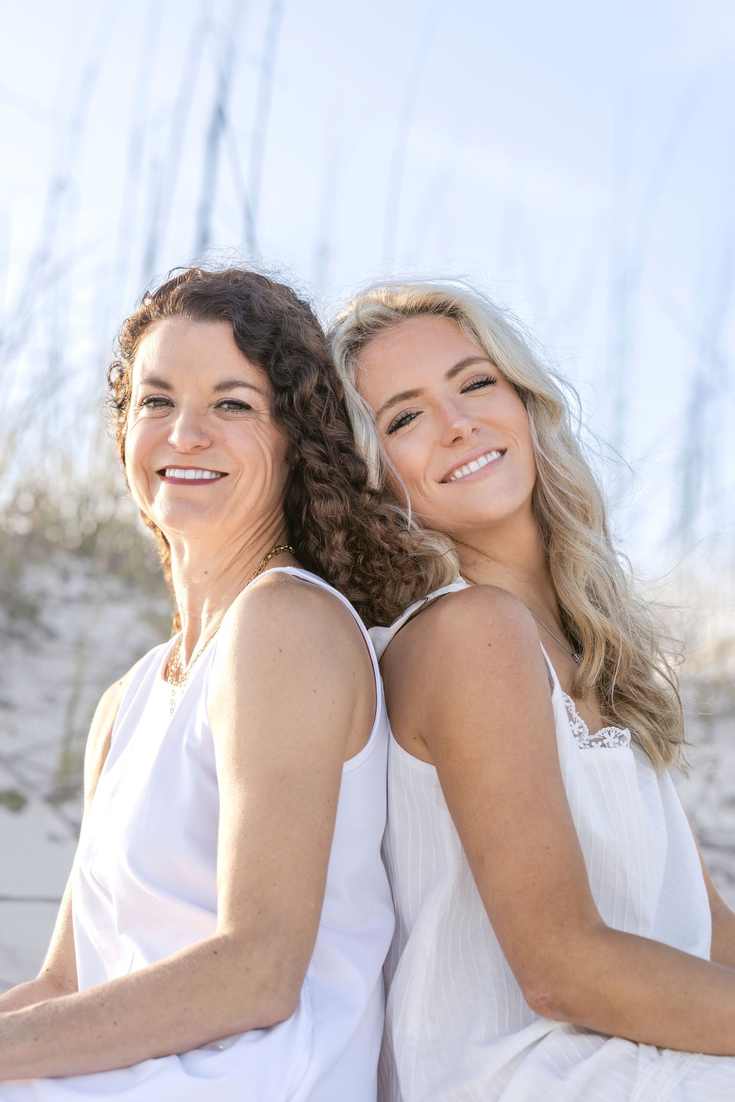 Two smiling women in white dresses pose back-to-back on a sunny beach with sandy dunes and tall grass behind them.