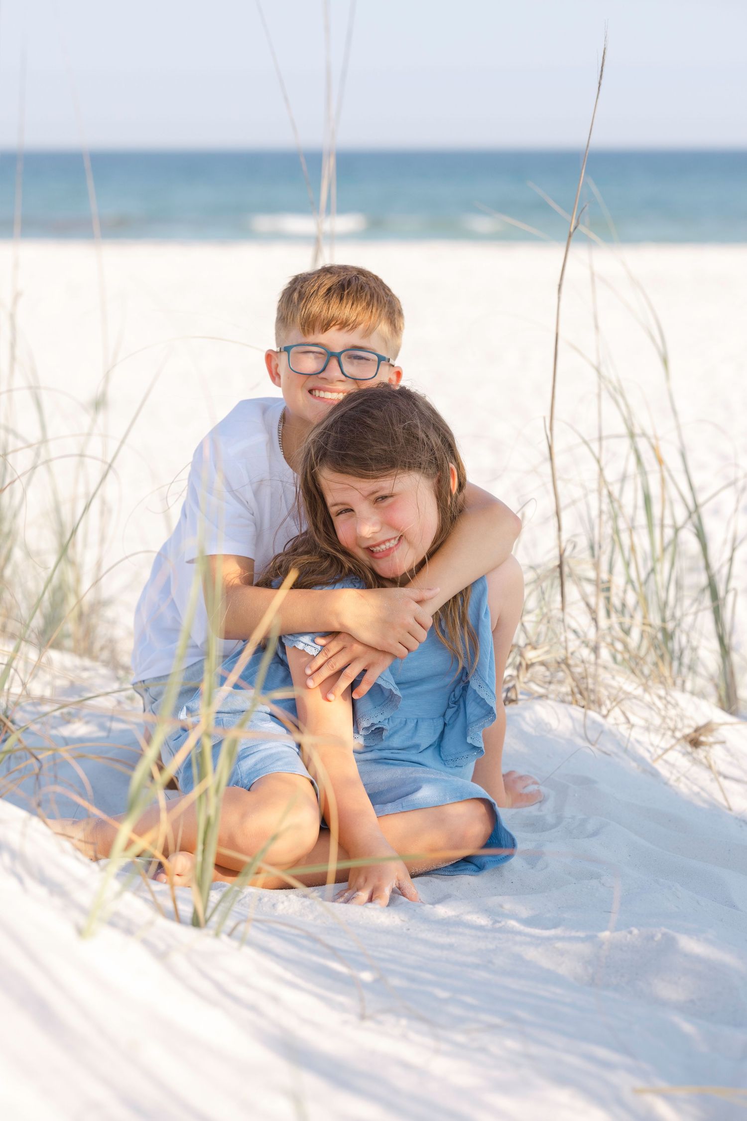 Two children sit together on white sand dunes at the beach, with the boy hugging the girl from behind.