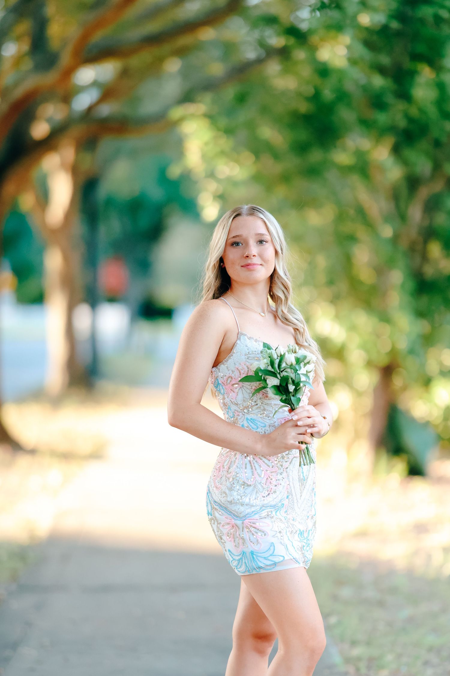 Young blonde woman in floral mini dress holding green bouquet, posing on tree-lined pathway outdoors.