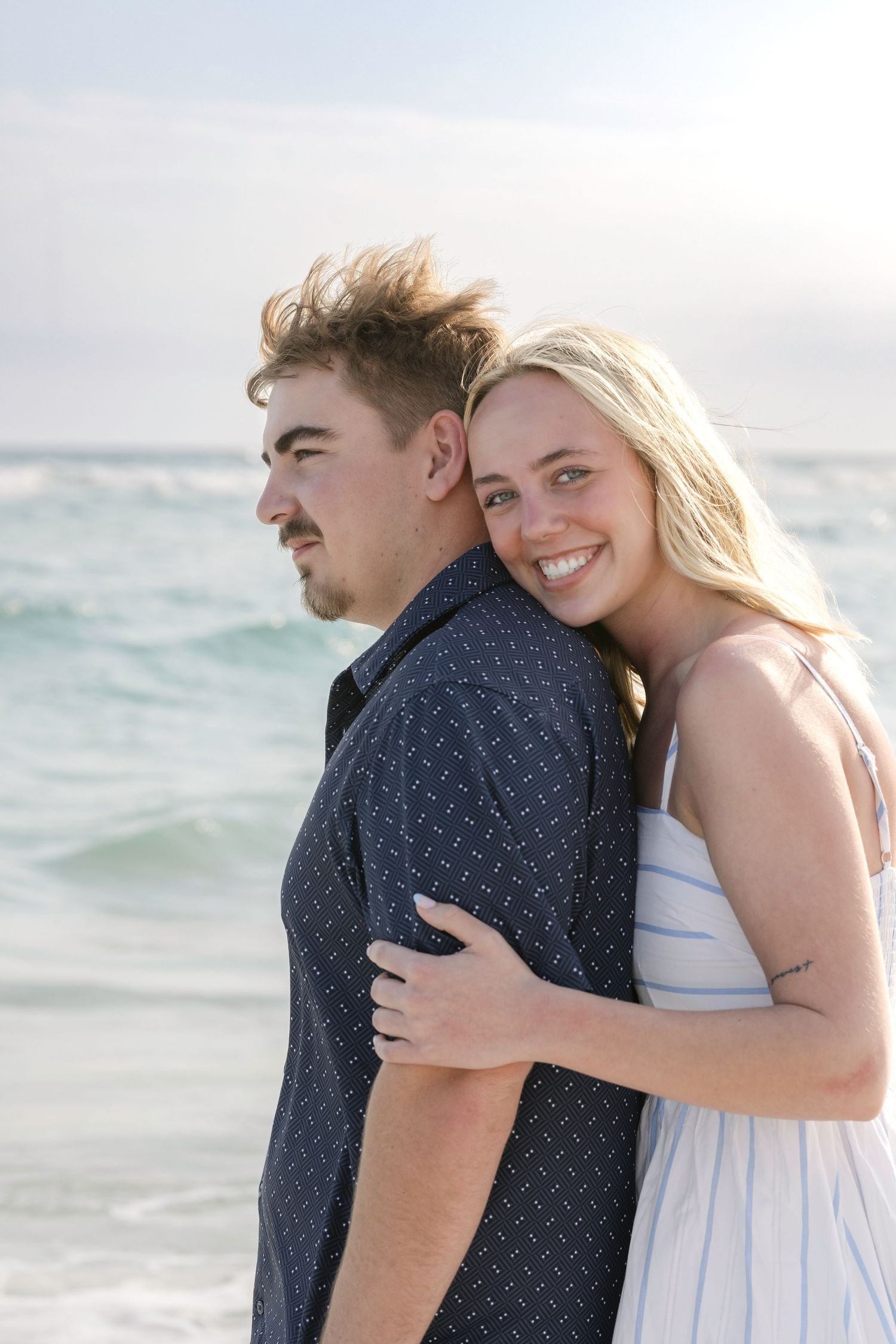 Happy blonde woman hugging man from behind on sunny beach with turquoise ocean waves.