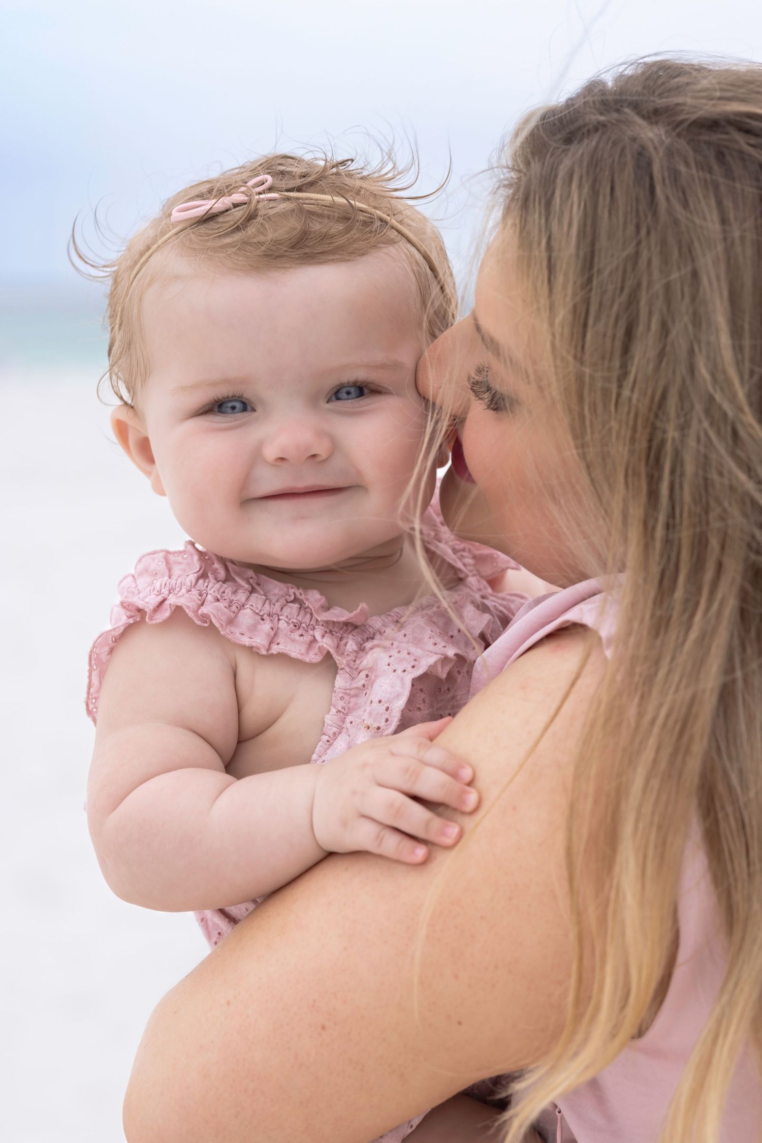 Mother kissing her smiling baby girl in a pink dress on a sunny beach.