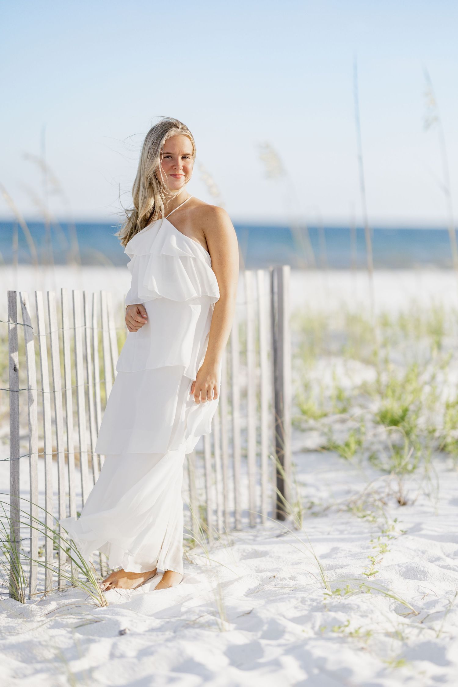 A blonde woman in a white one-shoulder maxi dress poses gracefully by a beach fence on white sand dunes.