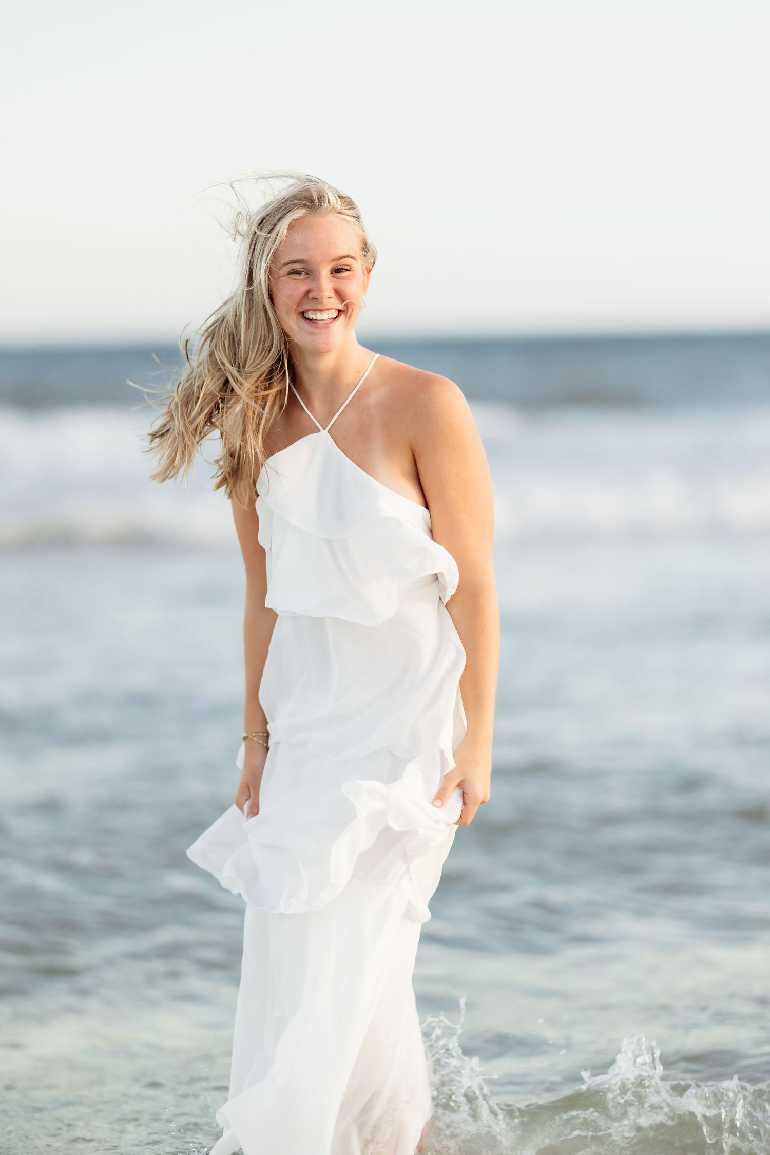 Smiling blonde woman in white ruffled dress standing in ocean waves on sunny beach.