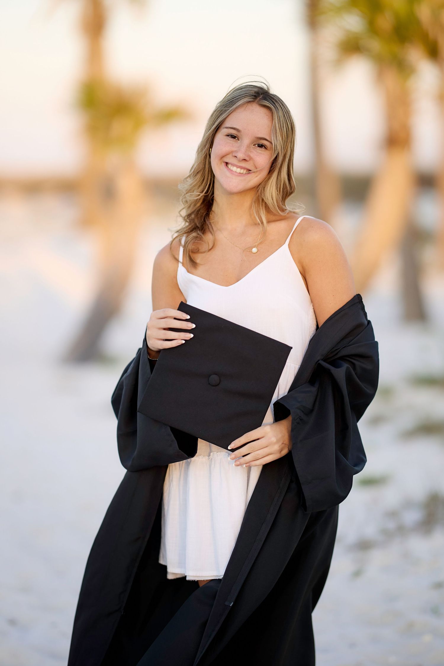 A person wearing a black cardigan over a white top poses outdoors with a soft golden sunset glow in the background.