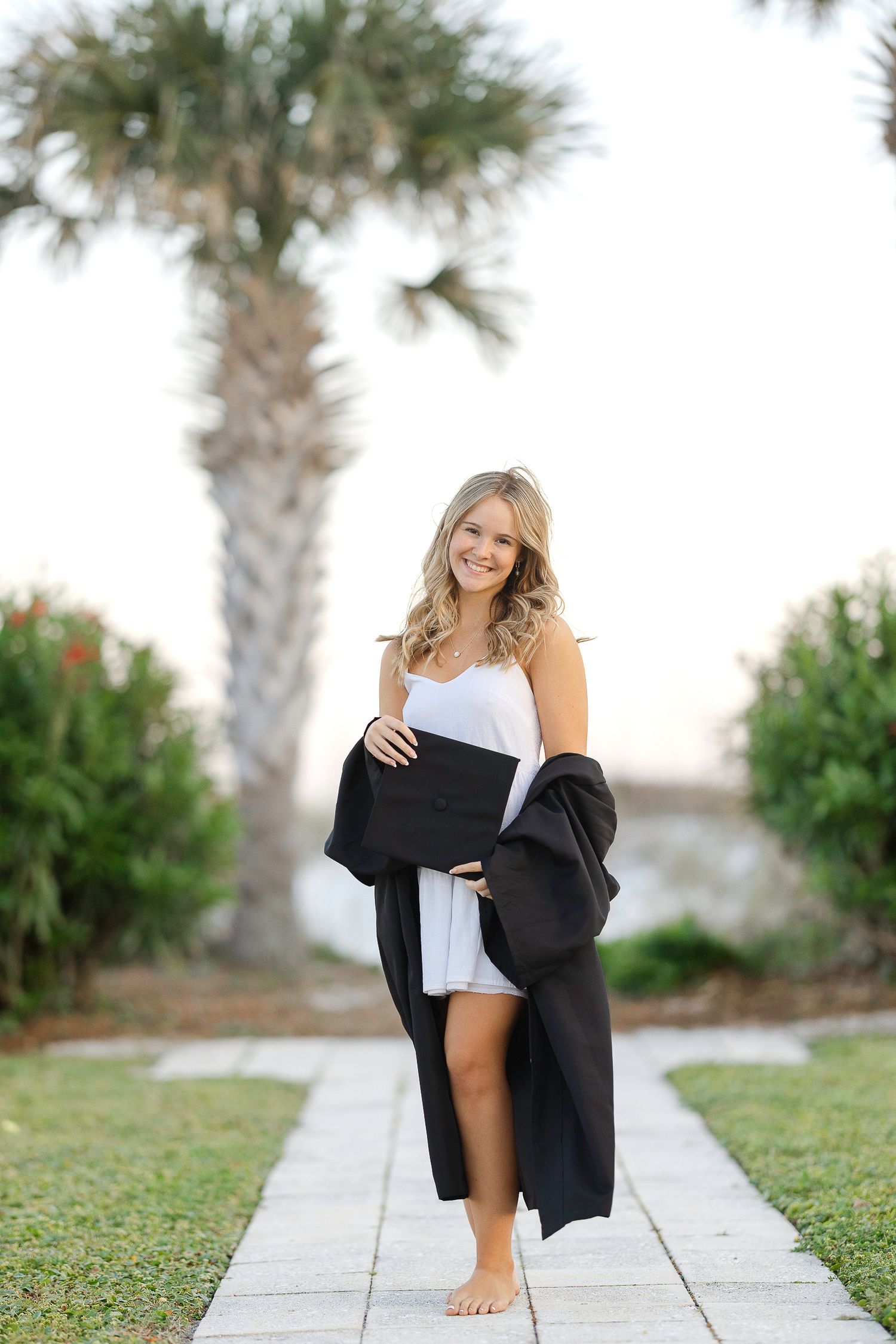 A graduate sits on a stone path surrounded by palm trees and grass during a casual outdoor photo session.