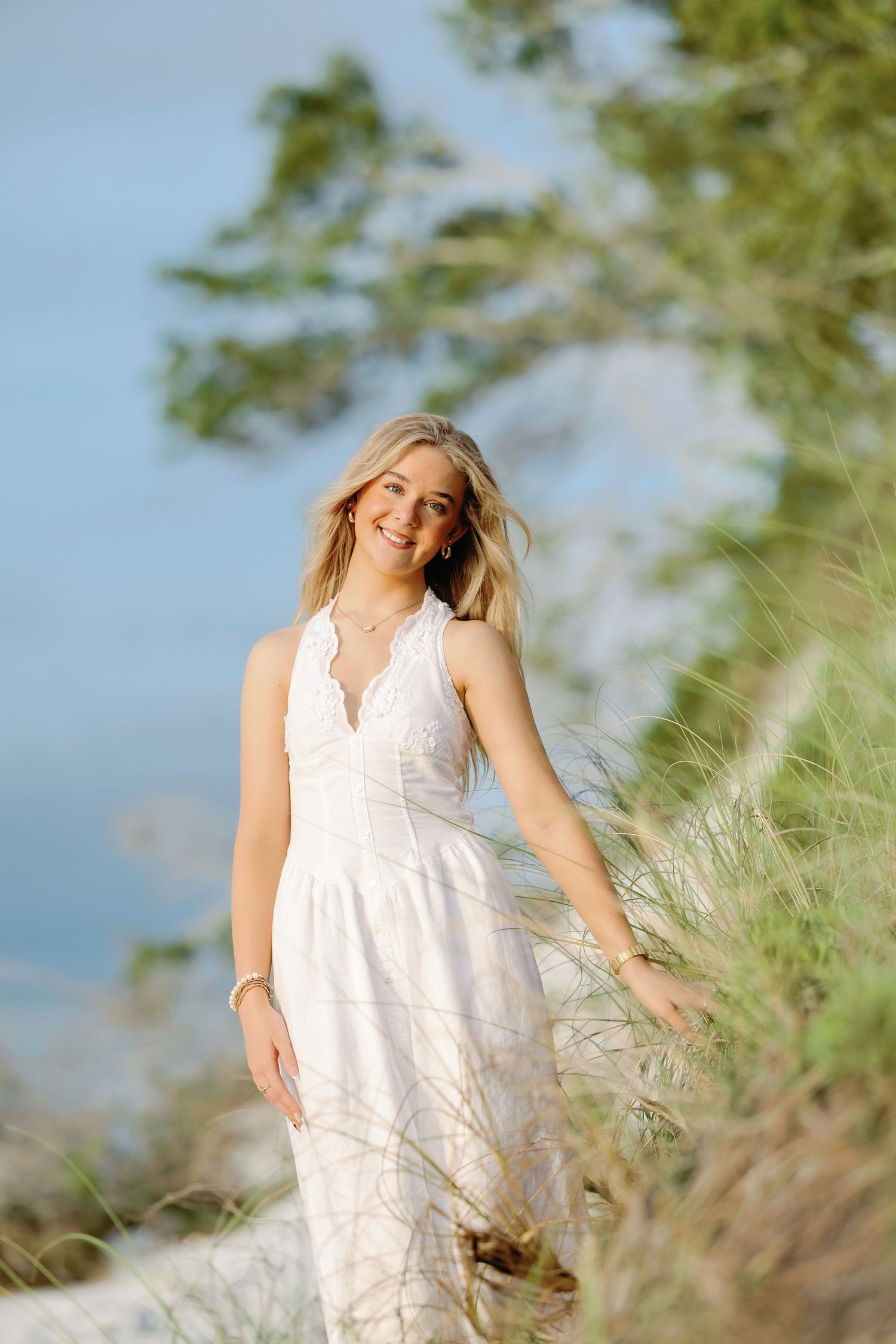 A person in a flowing white lace dress poses on a hillside with sunlit foliage and blue sky in the background.