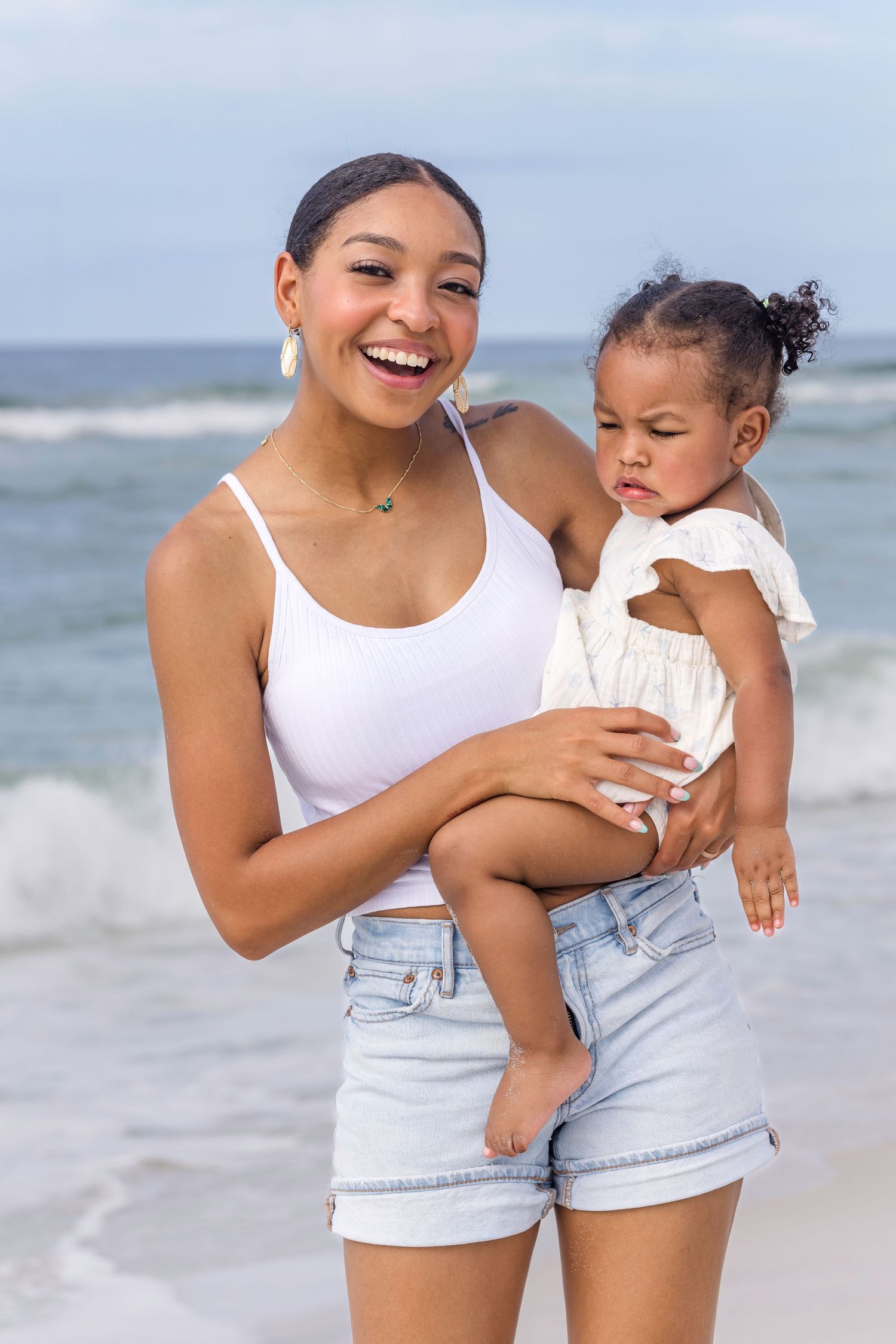 Smiling young Black woman holds a toddler girl on a sunny beach with ocean waves.