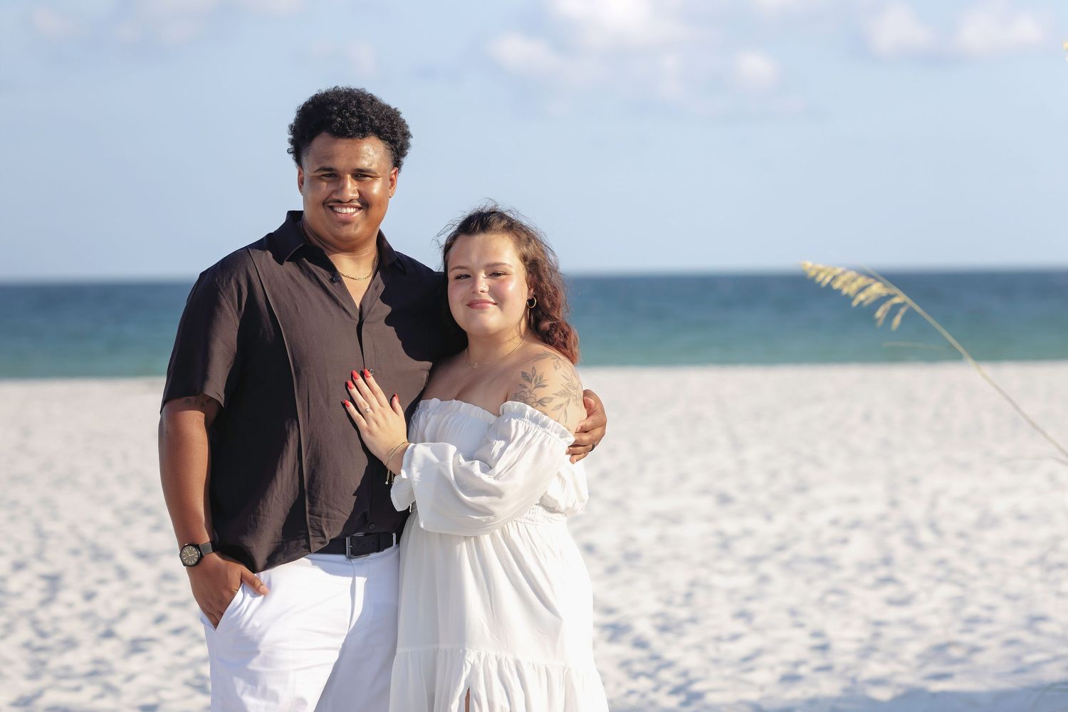 Couple posing together on a white sandy beach, man in black shirt and woman in white off-shoulder dress.