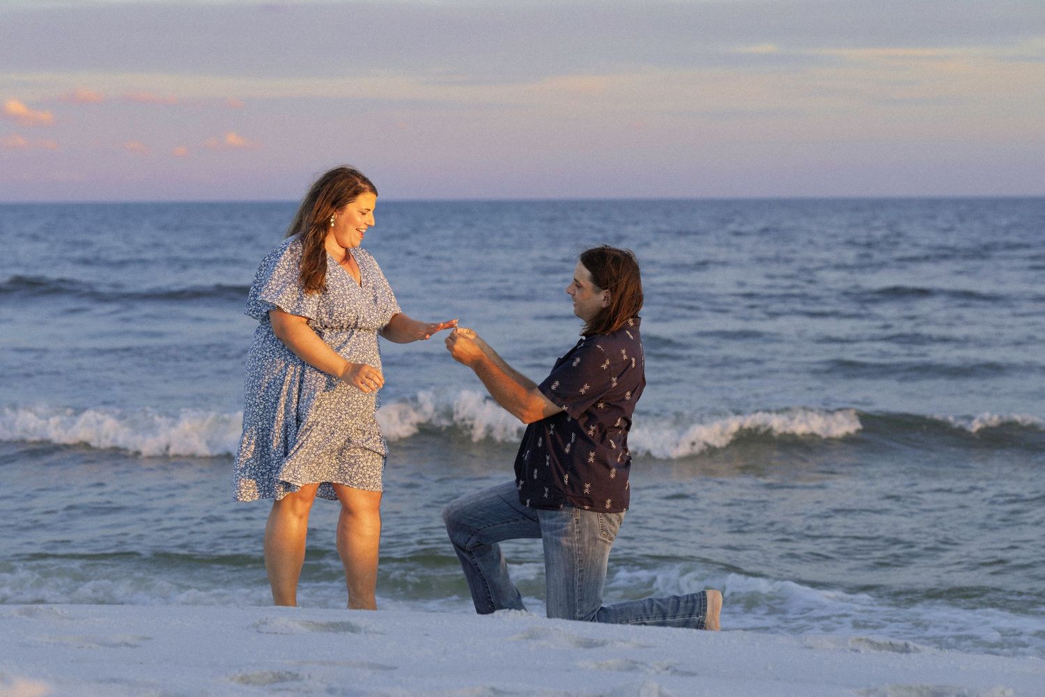 Man proposes to pregnant woman on a beach at sunset, kneeling in shallow ocean waves holding her hand.