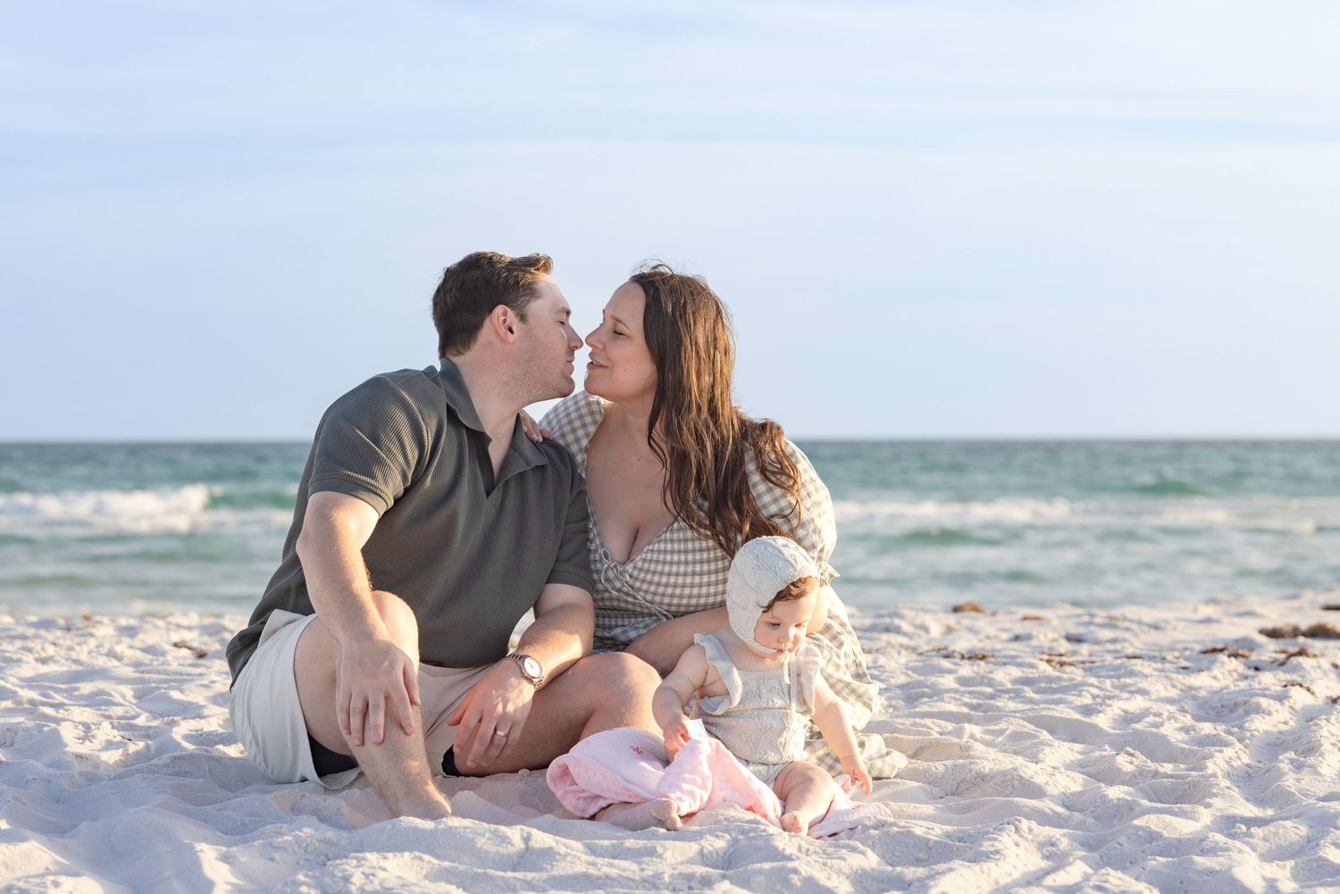 Couple sharing a romantic moment on a white sand beach with their baby sitting in front of them.